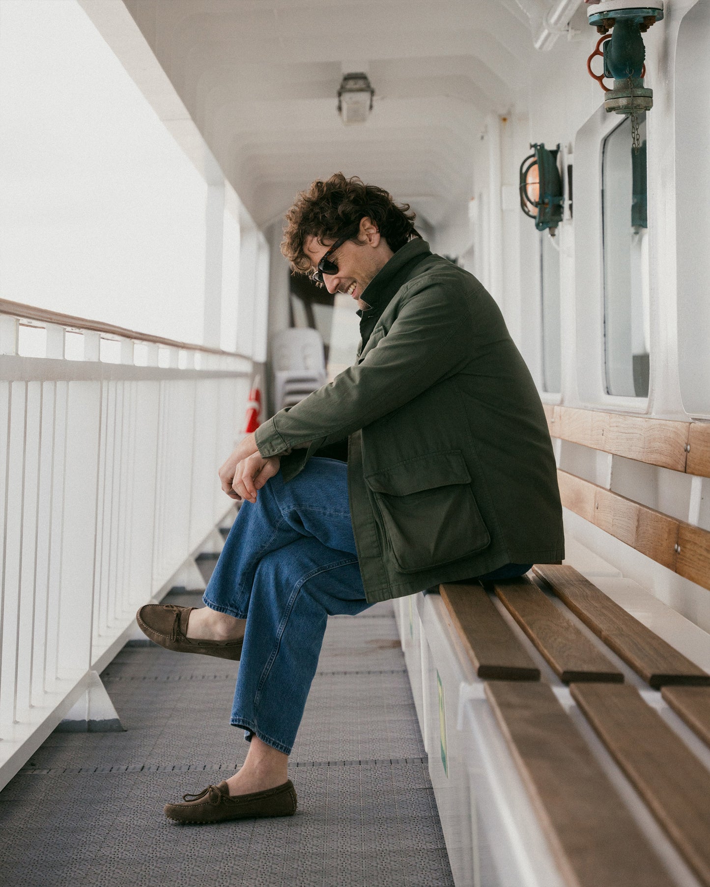 A man wearing a green jacket and blue jeans with Tofta – Taupe Suede driving shoes, sitting on a bench on a boat deck.