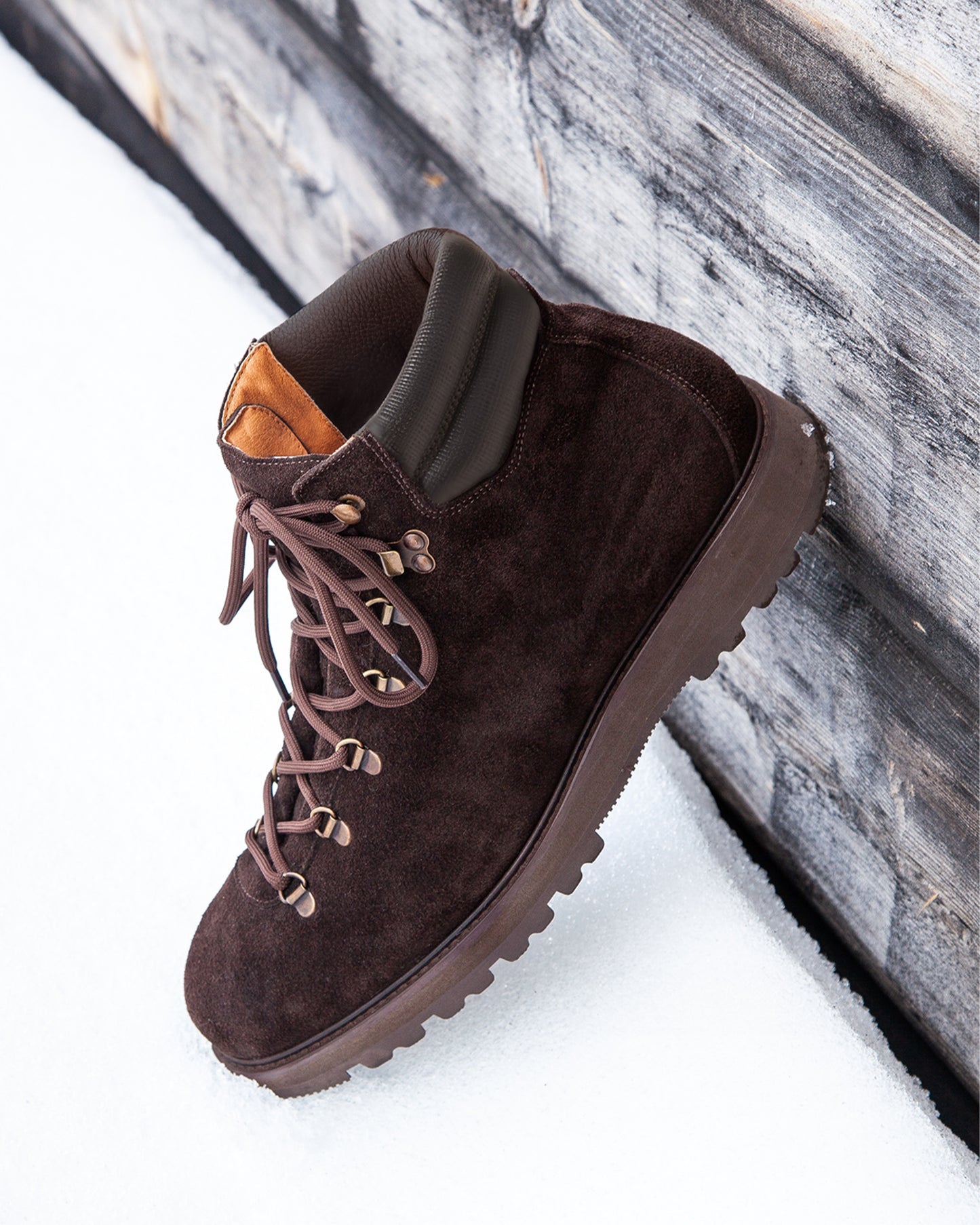A Duved II – Dark Brown Suede boot resting in the snow against a weathered wooden wall.