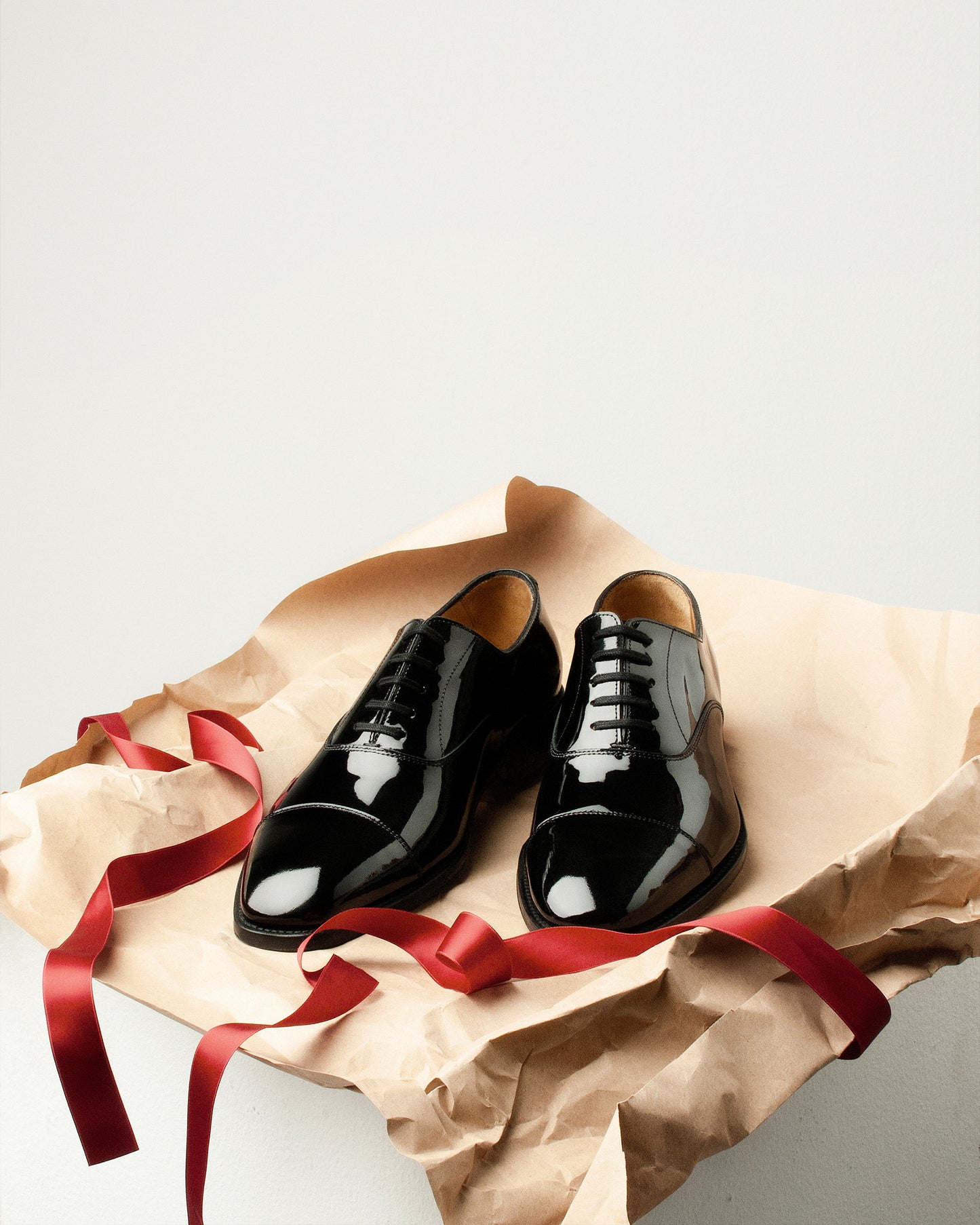 A pair of black patent leather Oxford shoes on crumpled brown paper with a red ribbon.
