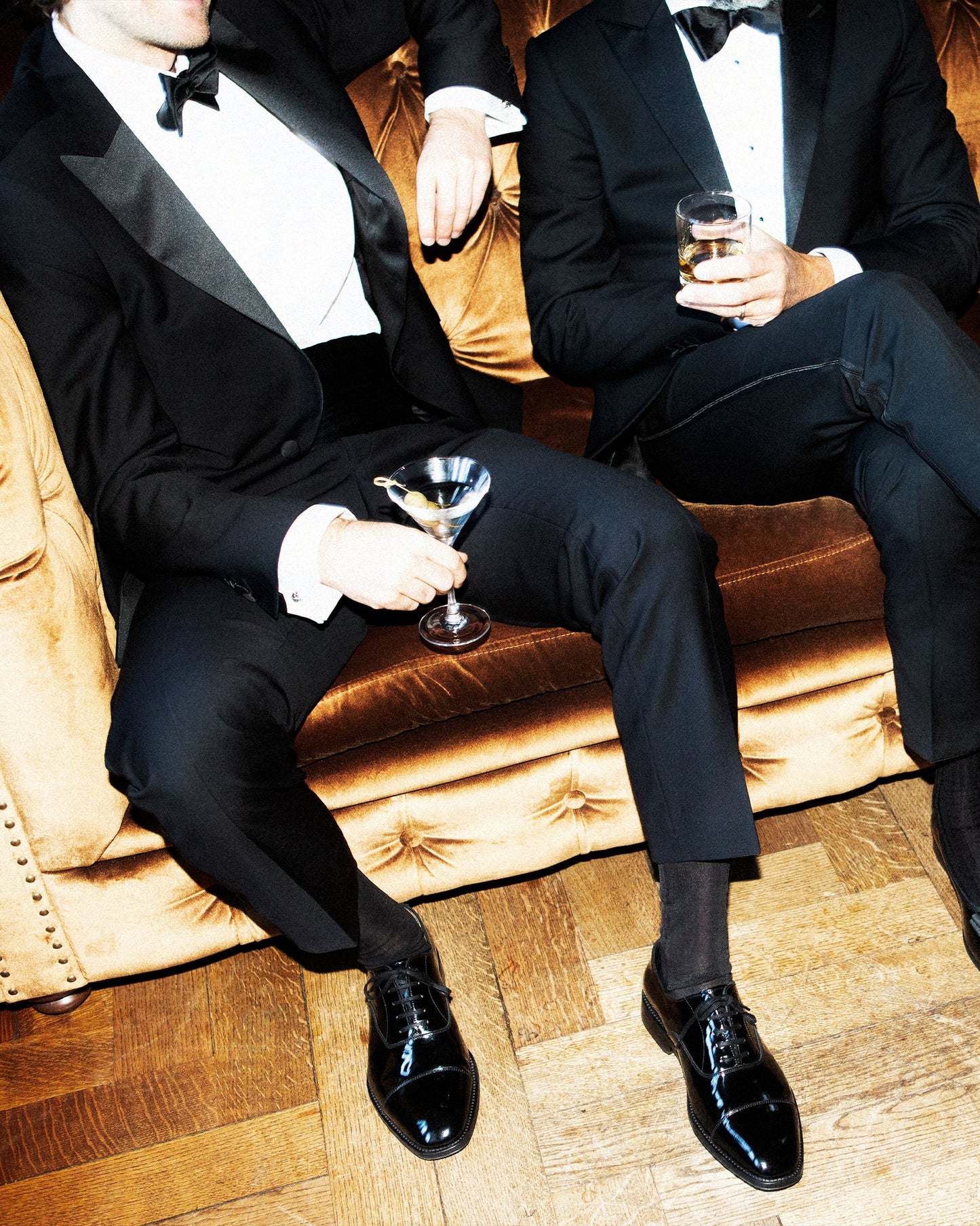 Two men in black tuxedos seated on a velvet sofa, each wearing Vinterviken II oxford shoes in black patent leather, their formal footwear gleaming and perfectly complementing their evening attire at a sophisticated event.
