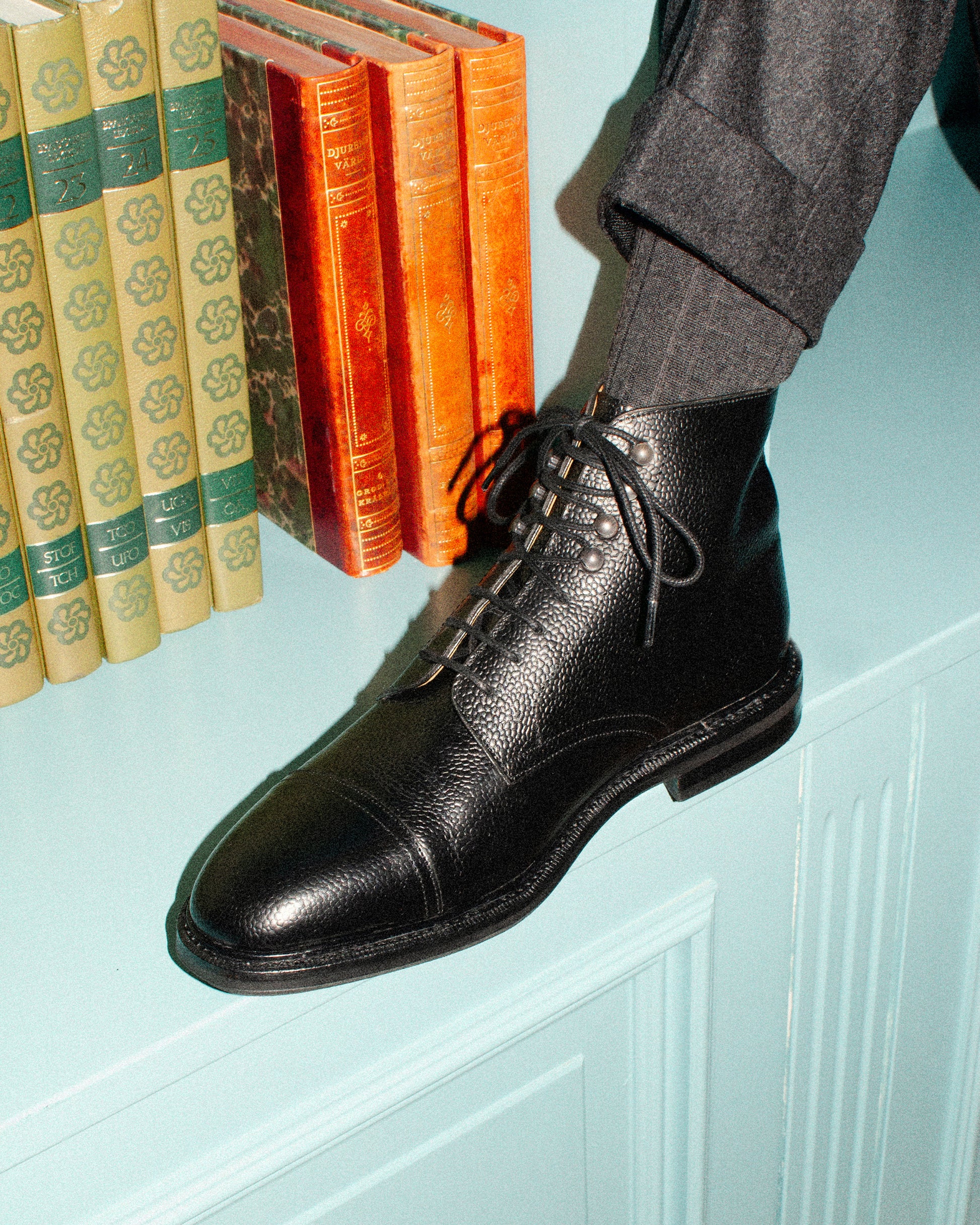 Close-up on a person wearing Abisko – Black Country Calf boots with grey trousers, resting by a row of books.