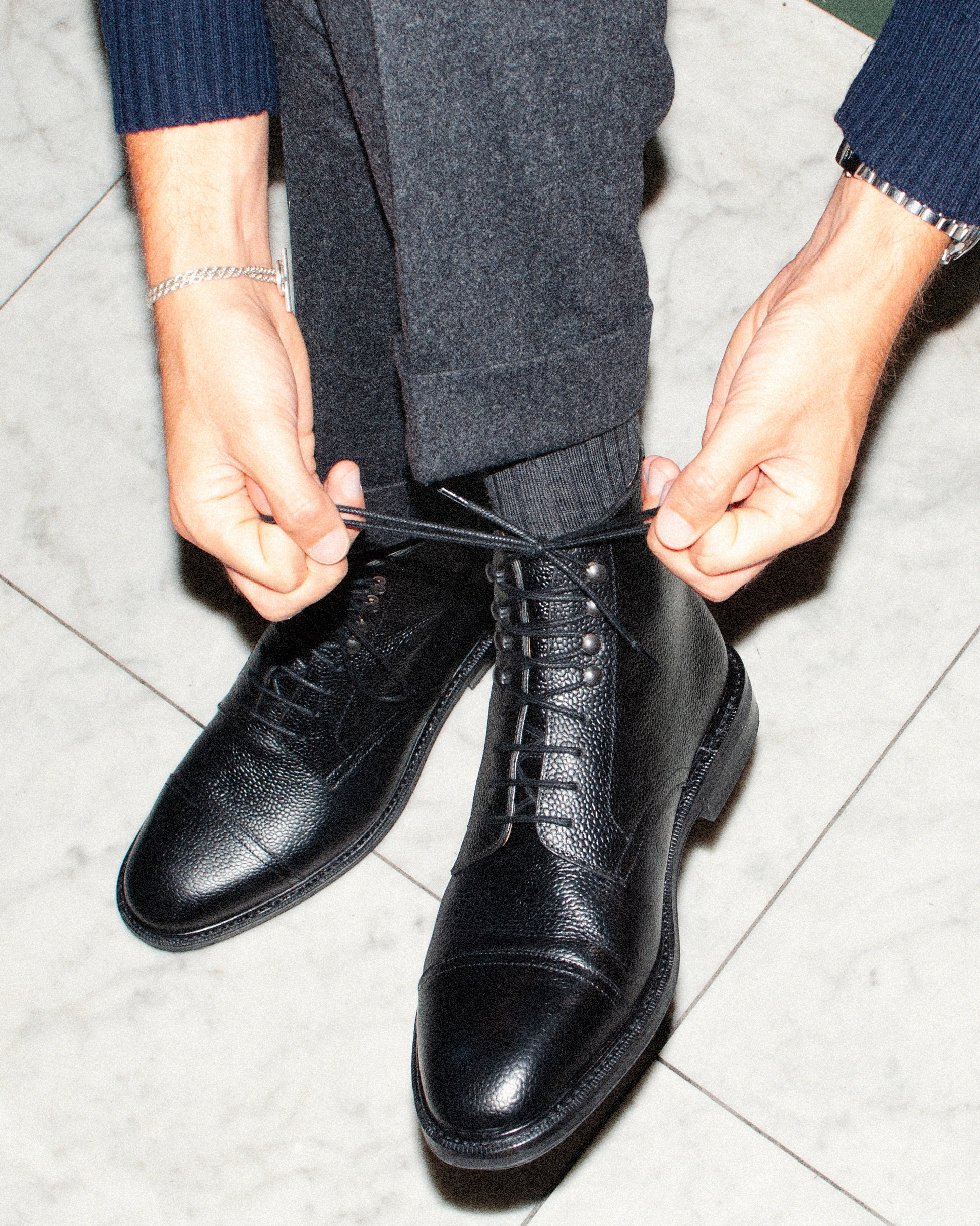 A close-up of a person tying the laces of their Abisko – Black Country Calf boots, worn with grey wool trousers.