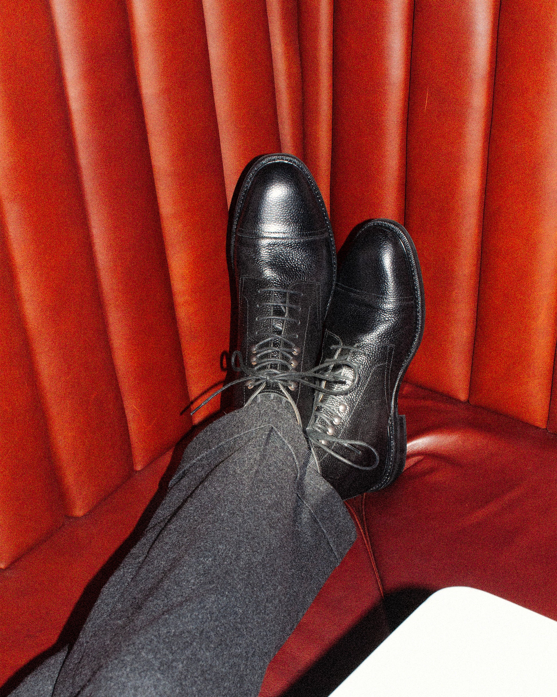 A person wearing Abisko – Black Country Calf boots with grey wool trousers, relaxing with feet up on a red leather seat.
