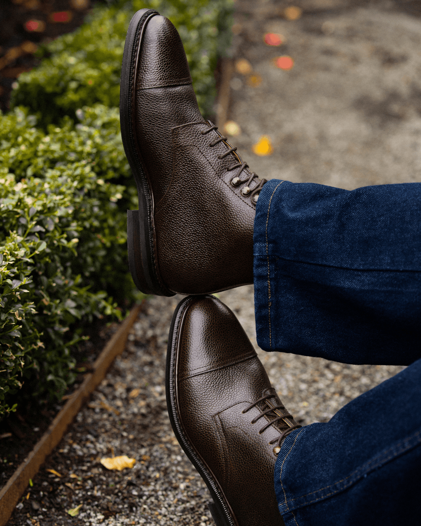 Close-up of a person wearing Abisko – Dark Brown Country Calf boots with blue jeans, with feet crossed outdoors.
