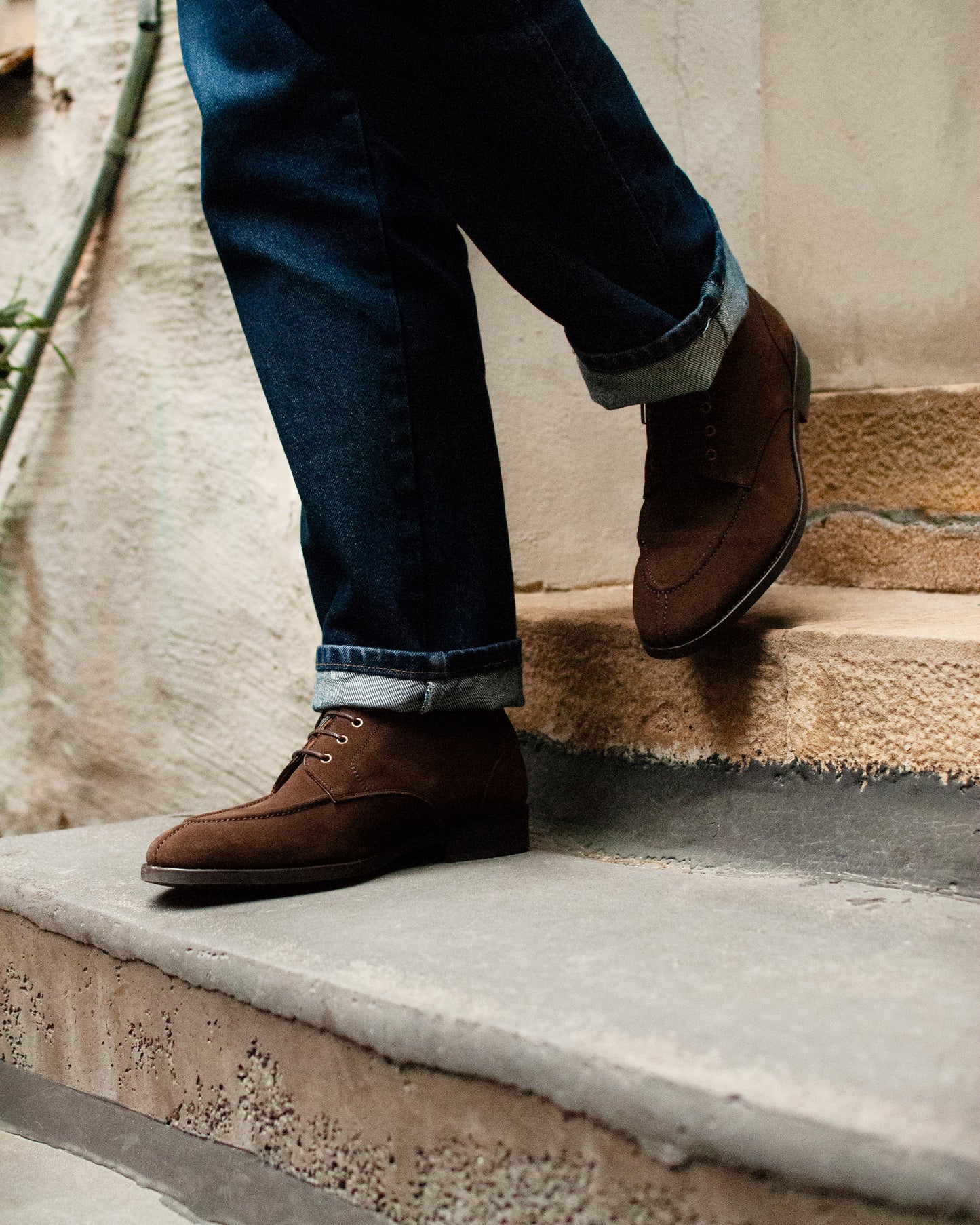 A person wearing Gothem dark brown suede boots and cuffed jeans, walking down a set of stone stairs.