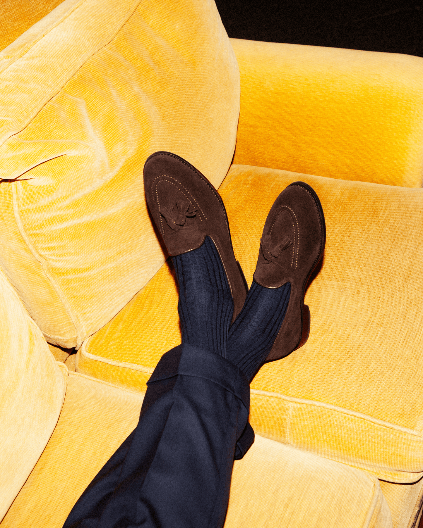 Person wearing Mölle II – Dark Brown Suede loafers and navy trousers, resting feet on a bright yellow sofa.