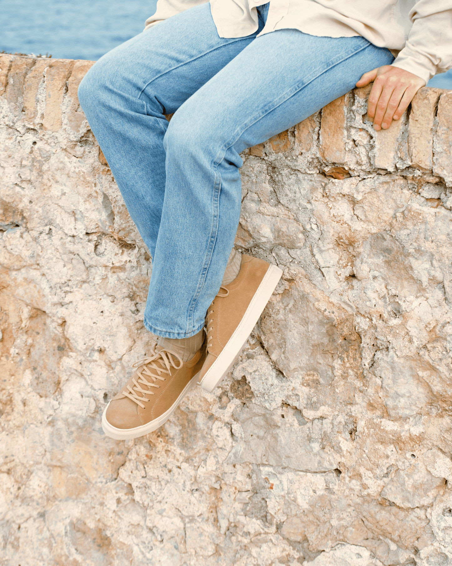A person sits on a stone wall by the sea, wearing light wash jeans and Oaxen – Beige Suede sneakers.