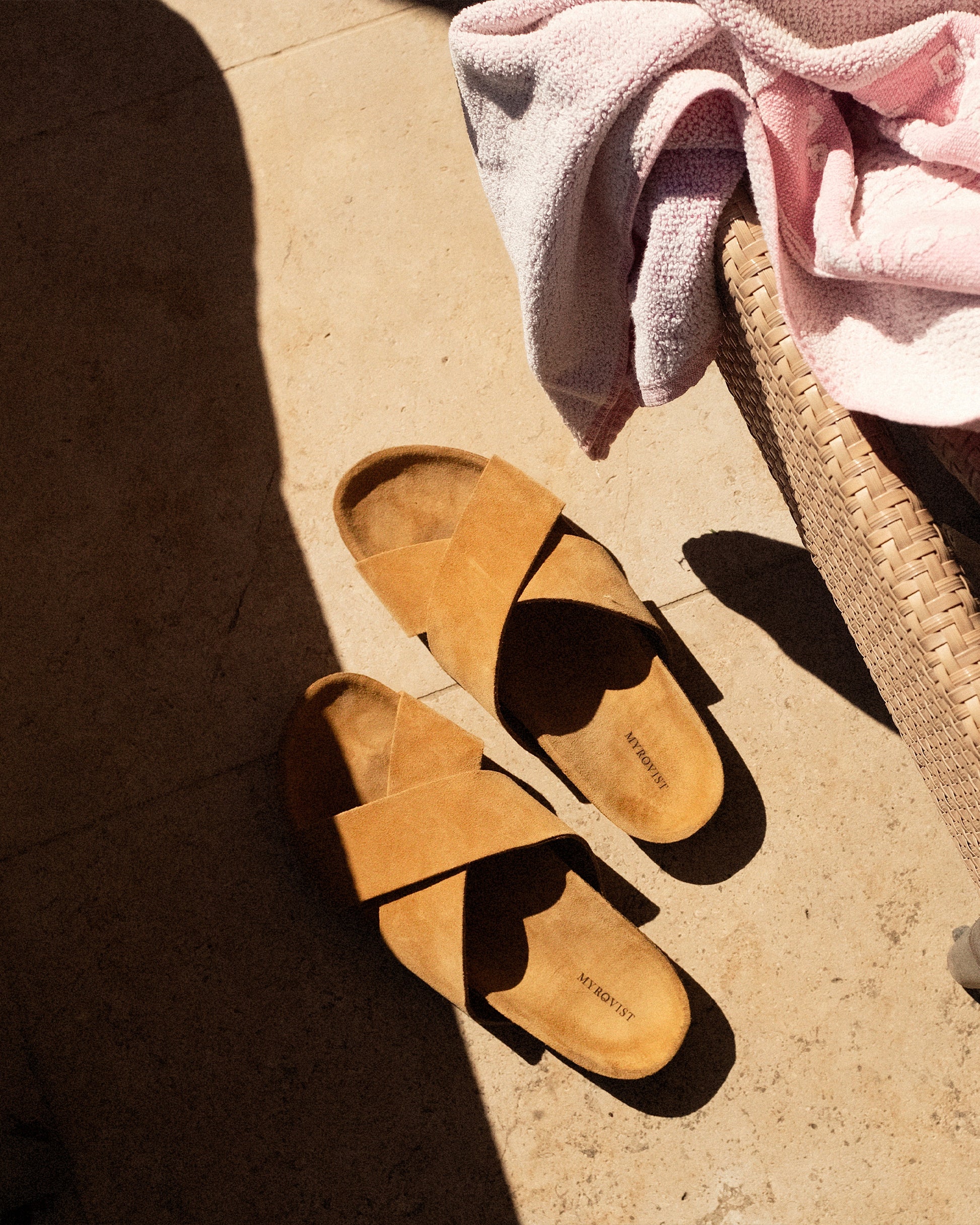 Overhead lifestyle shot of Solvik – Sand Suede sandals on a sunlit stone floor next to a wicker basket with a pink towel.