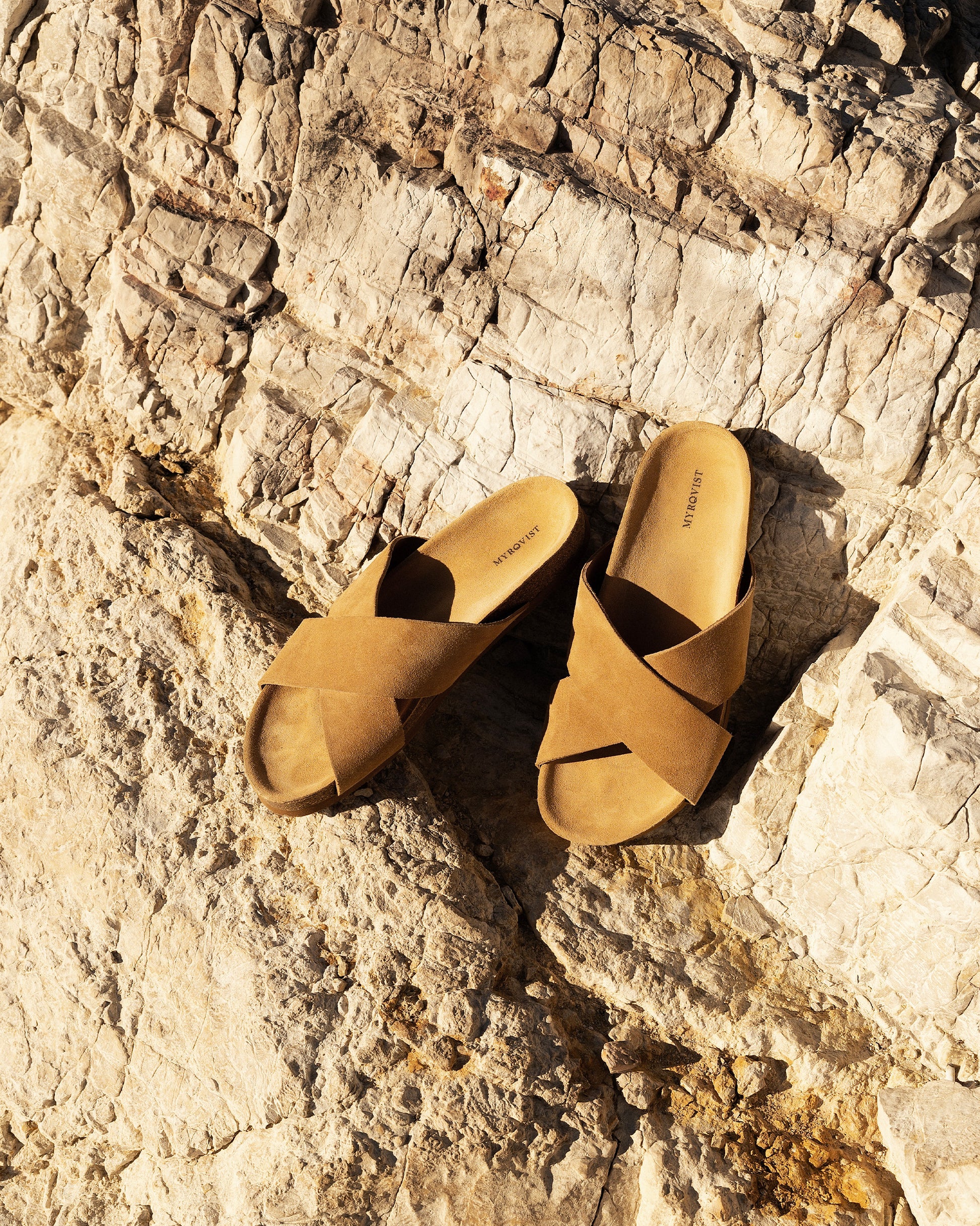 A pair of Solvik – Sand Suede sandals placed on a textured, sunlit rock surface.