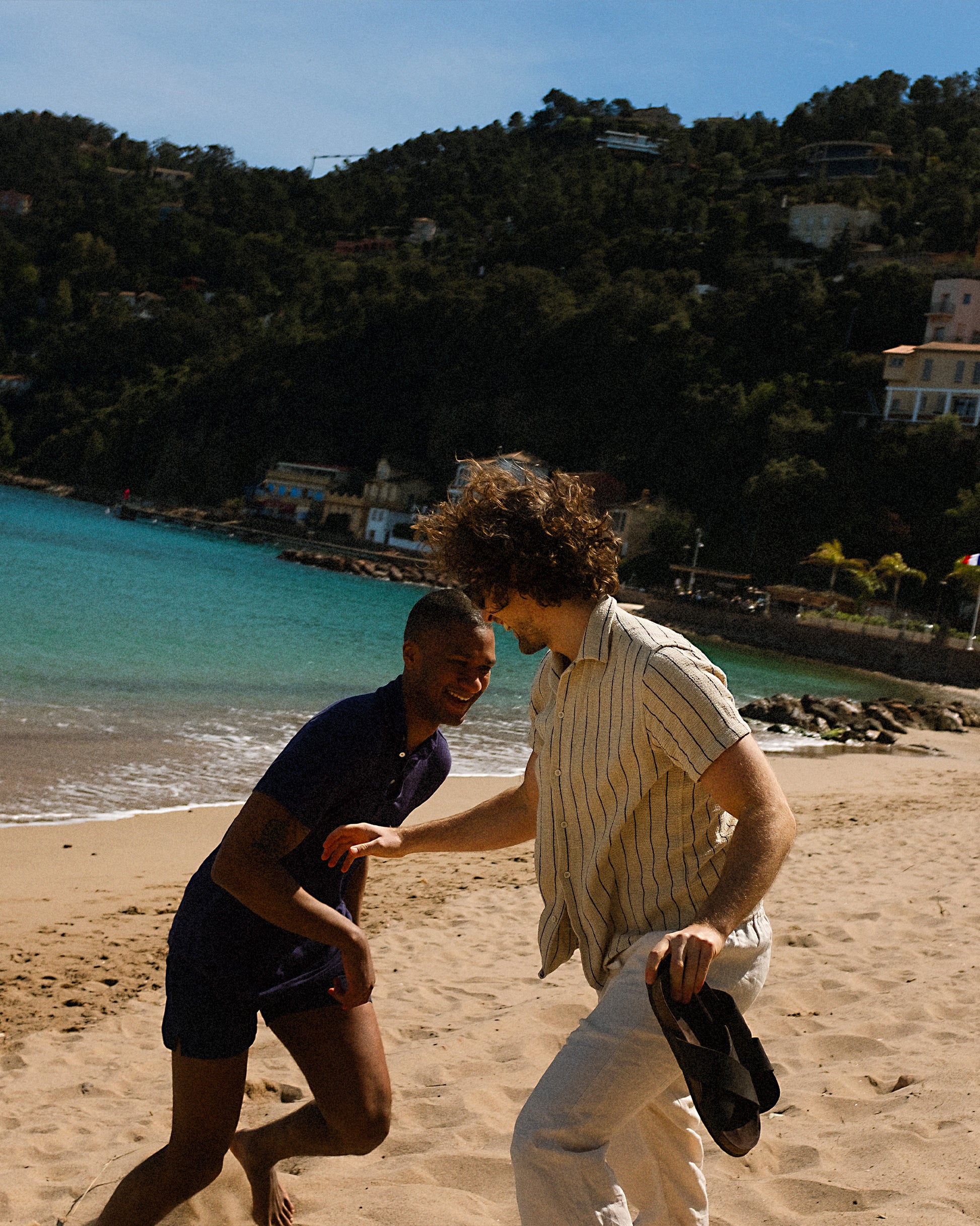 A man holding a pair of Solvik black suede sandals while laughing with a friend on a sunny beach.