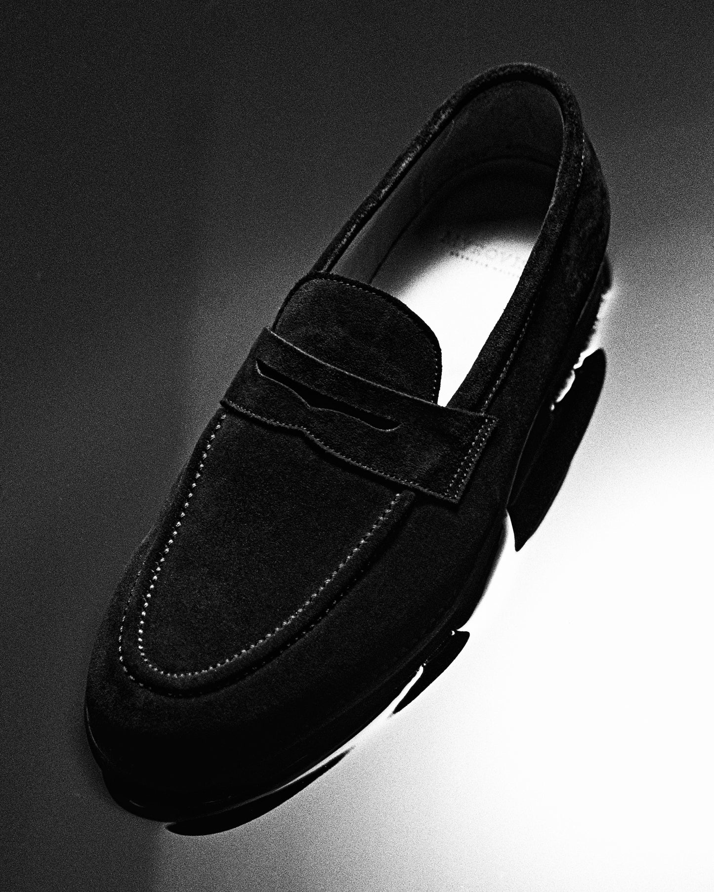 A black and white studio image of the Stenhammar – Black Suede loafer, shot from above against a dark, reflective backdrop.