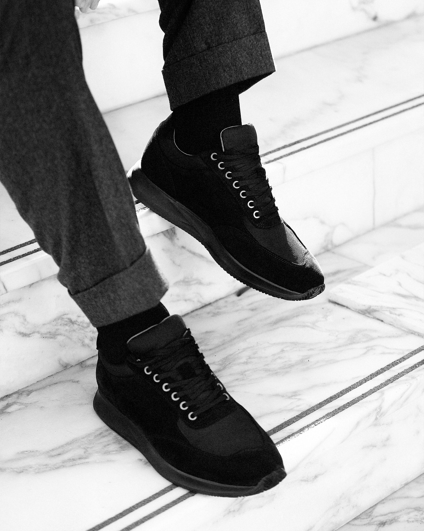 Close-up of Stensund – Black Suede trainers worn with cuffed grey trousers on a marble staircase.