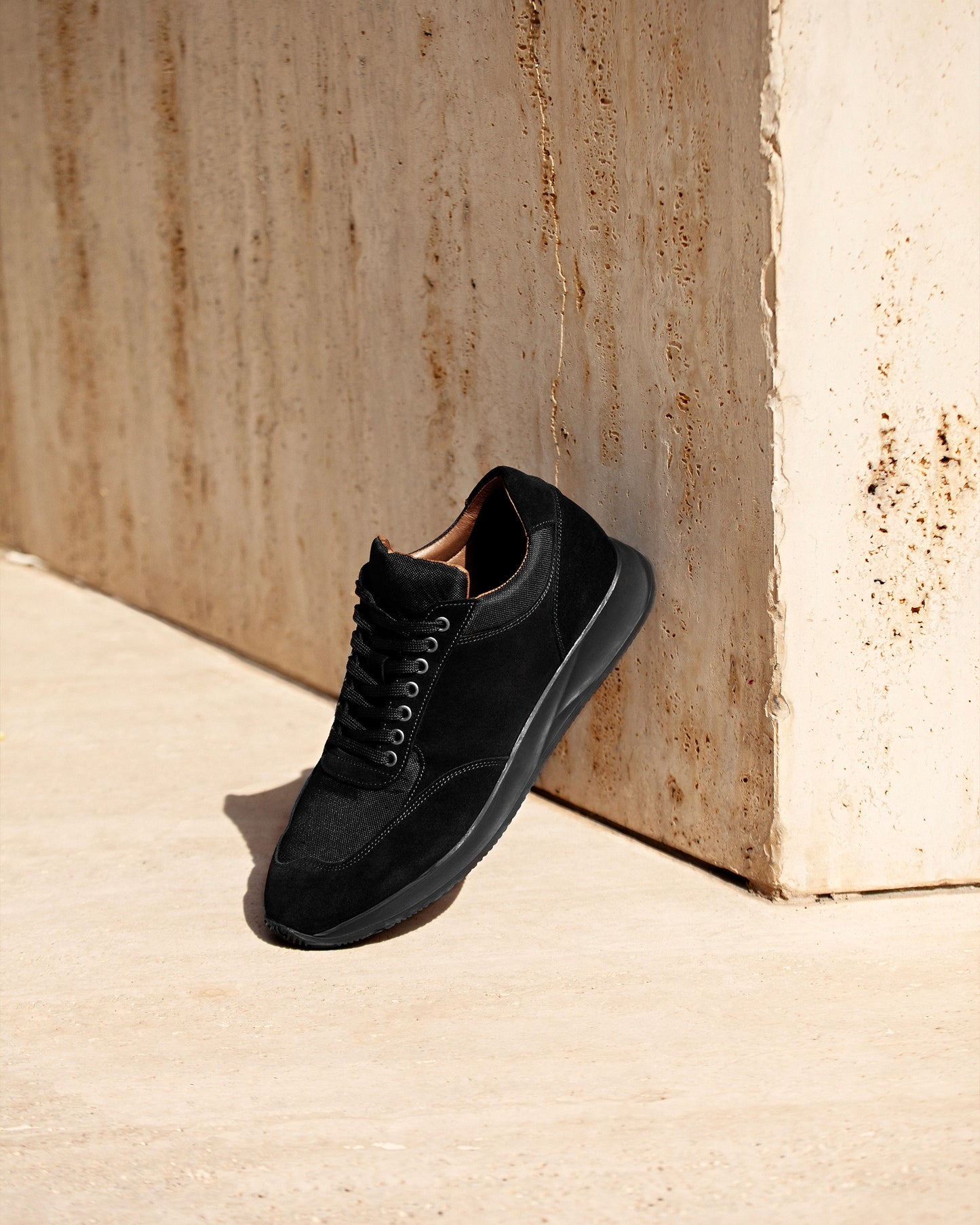 Stensund – Black Suede trainer leaning against a textured beige wall in bright sunlight.