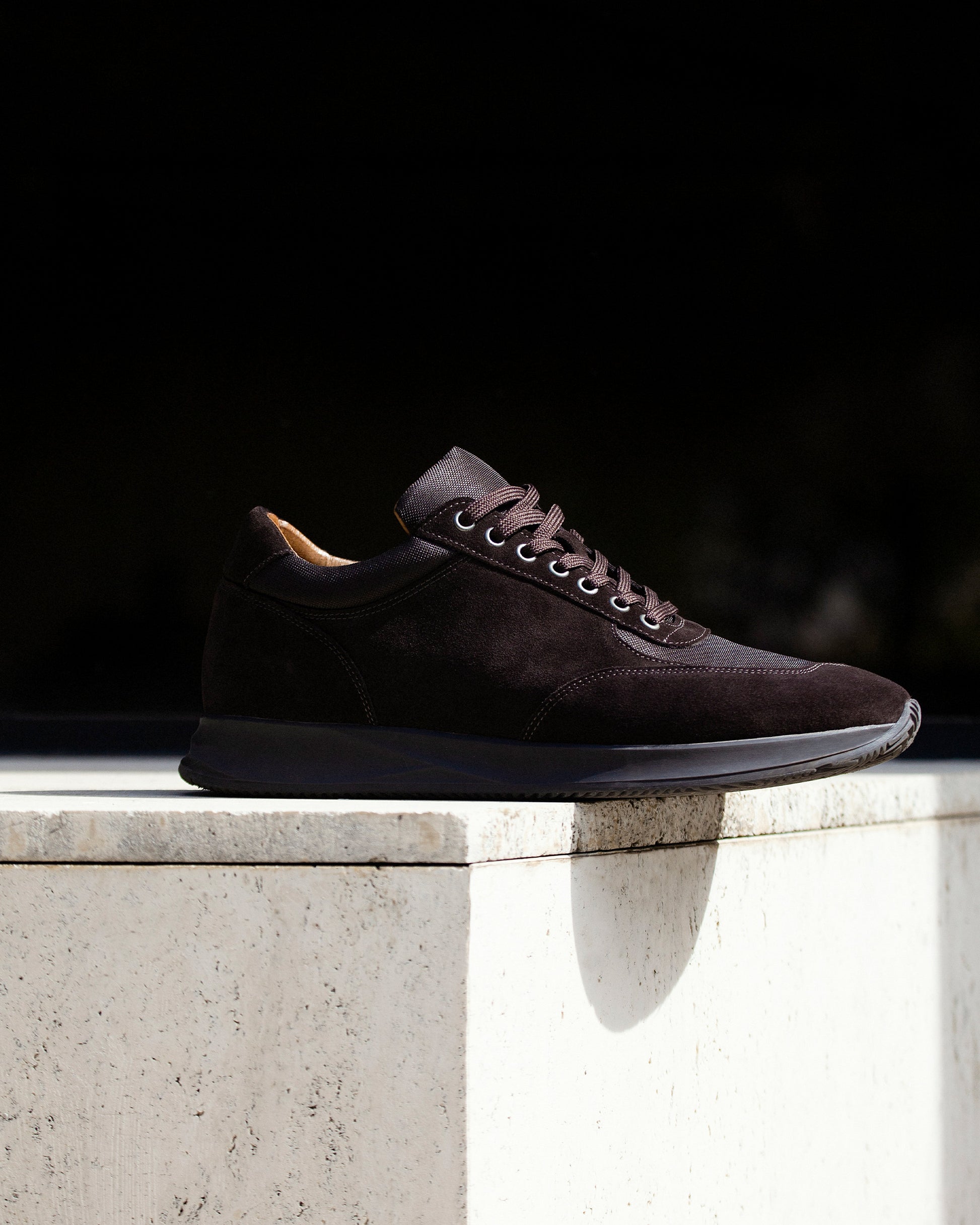 A close-up of Stensund – Dark Brown Suede sneaker, detailing its side profile as it rests on a sunlit stone ledge.