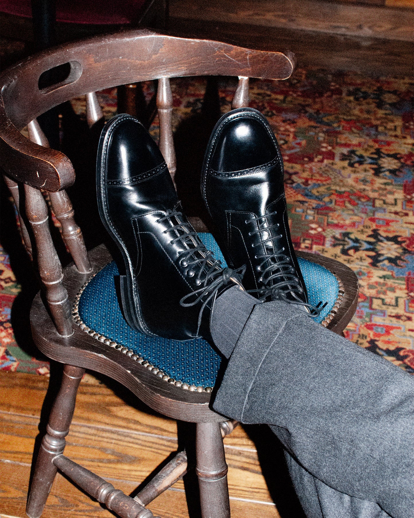 Close-up of a person wearing Tjärnö – Black Calf boots and grey trousers, with their feet up on a wooden chair.