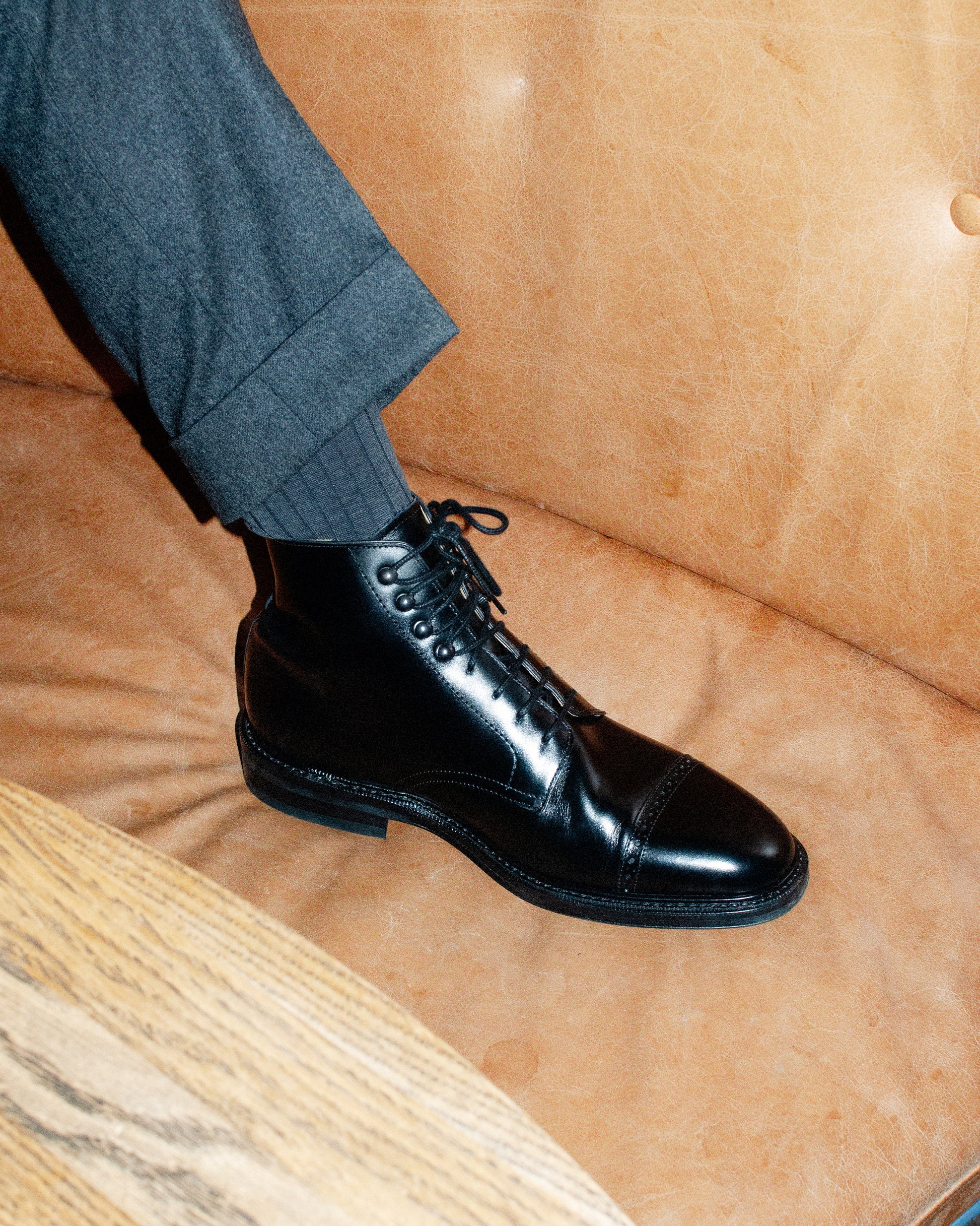 A person wearing Tjärnö – Black Calf boots with grey tailored trousers, seated on a tan leather sofa.