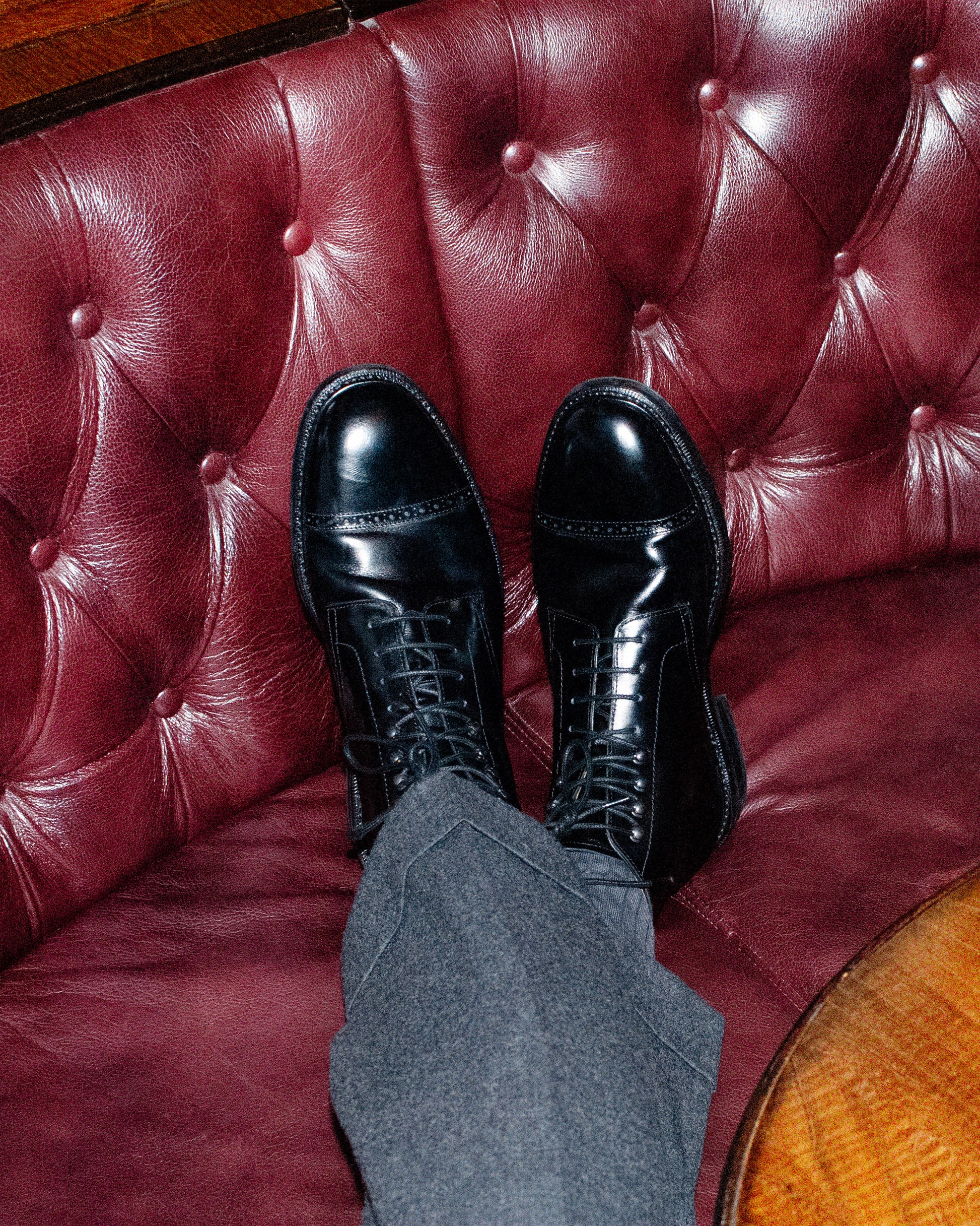 A person wearing Tjärnö – Black Calf boots and grey trousers, resting their feet on a tufted red leather sofa.