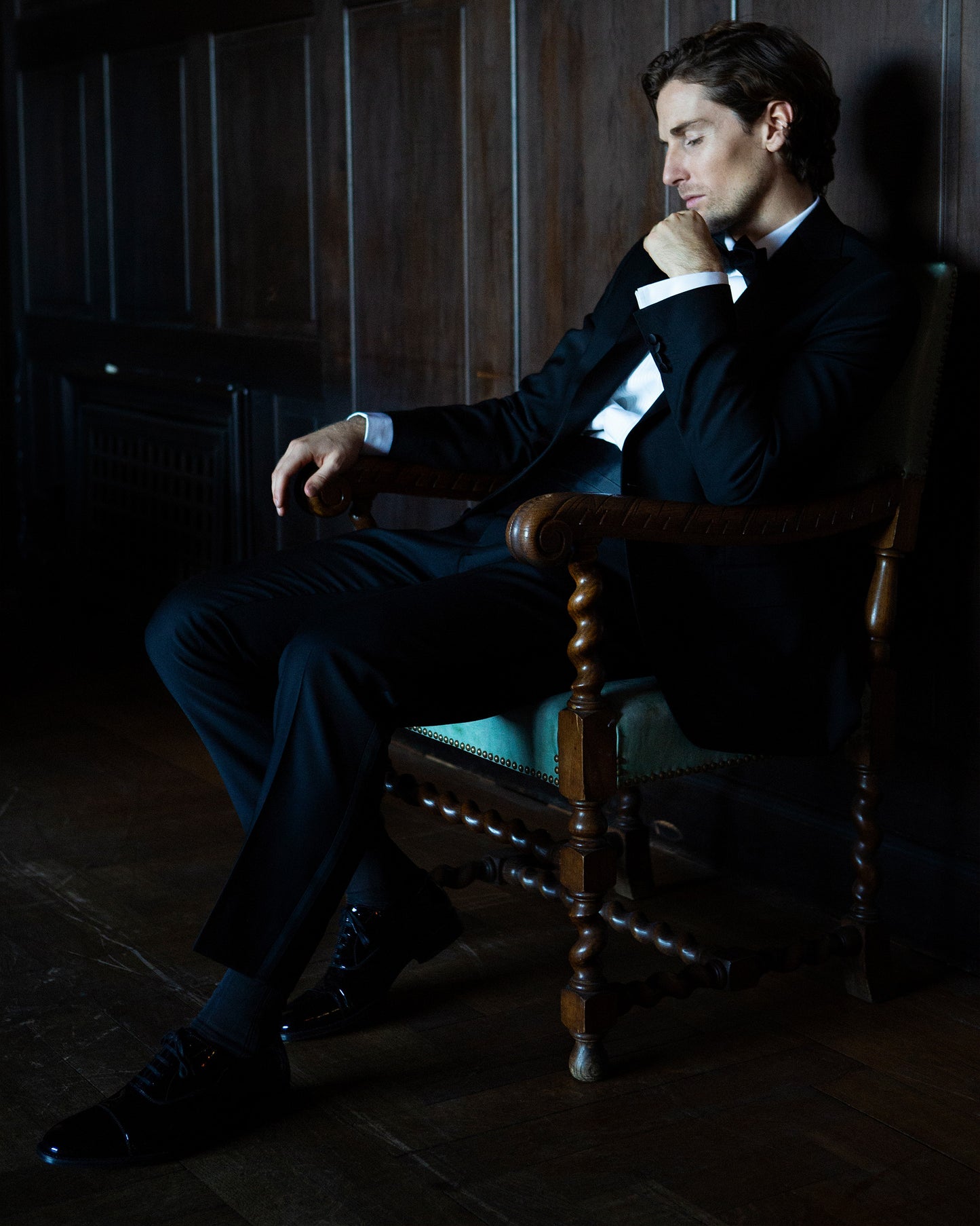 A man in a black tuxedo sits thoughtfully in an ornate wooden chair, wearing Vinterviken – Black Patent Leather shoes.