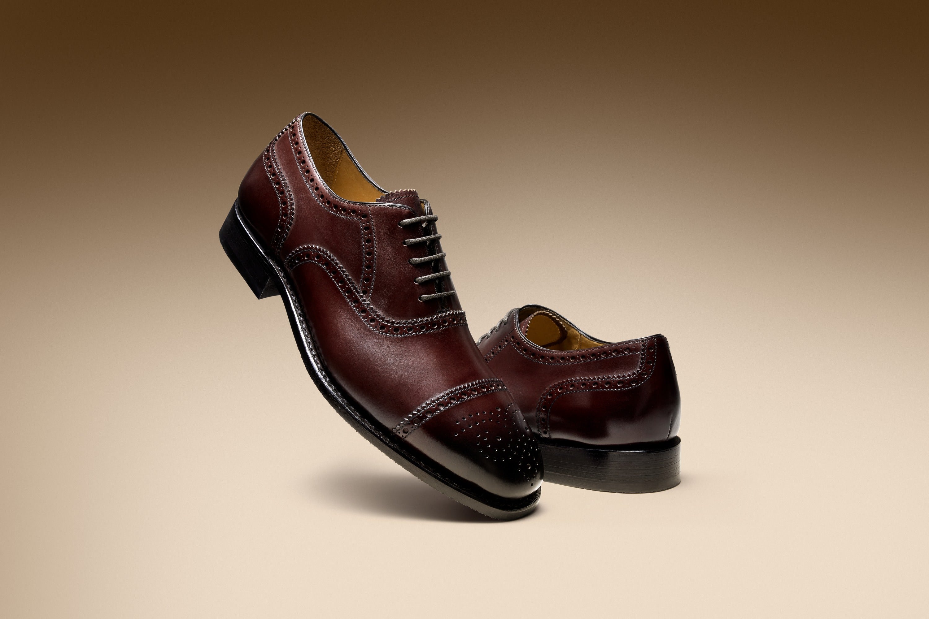 Two burgundy leather oxford shoes with brogue detailing, one leaning against the other in a warm studio setting.
