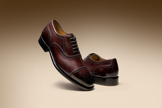 Two burgundy leather oxford shoes with brogue detailing, one leaning against the other in a warm studio setting.