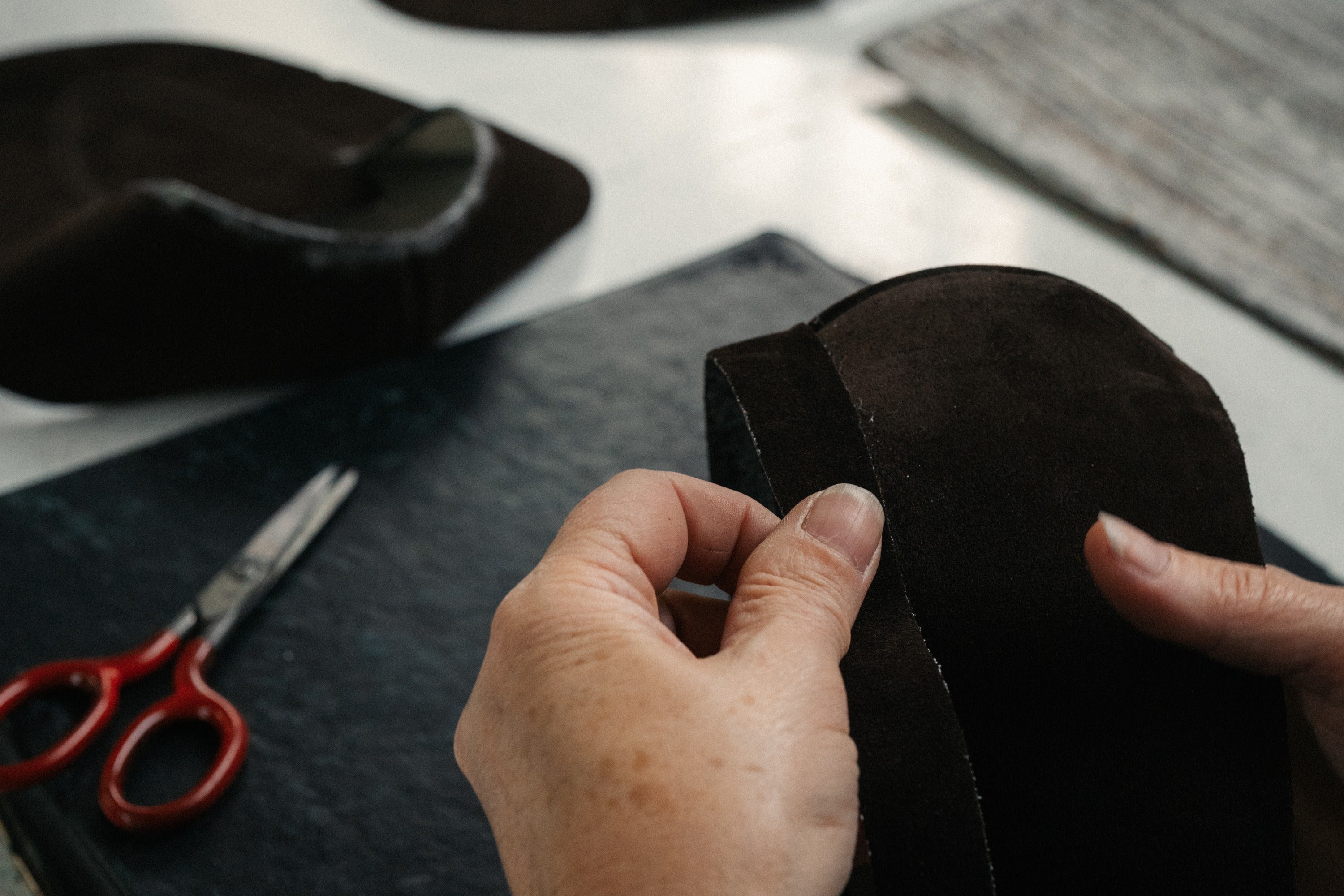 Hands stitch a dark suede shoe piece at a workbench, scissors resting beside.