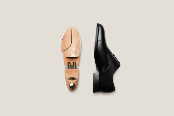 A cedar wooden shoe tree beside a black patent leather oxford, arranged at an angle on a pale linen surface.