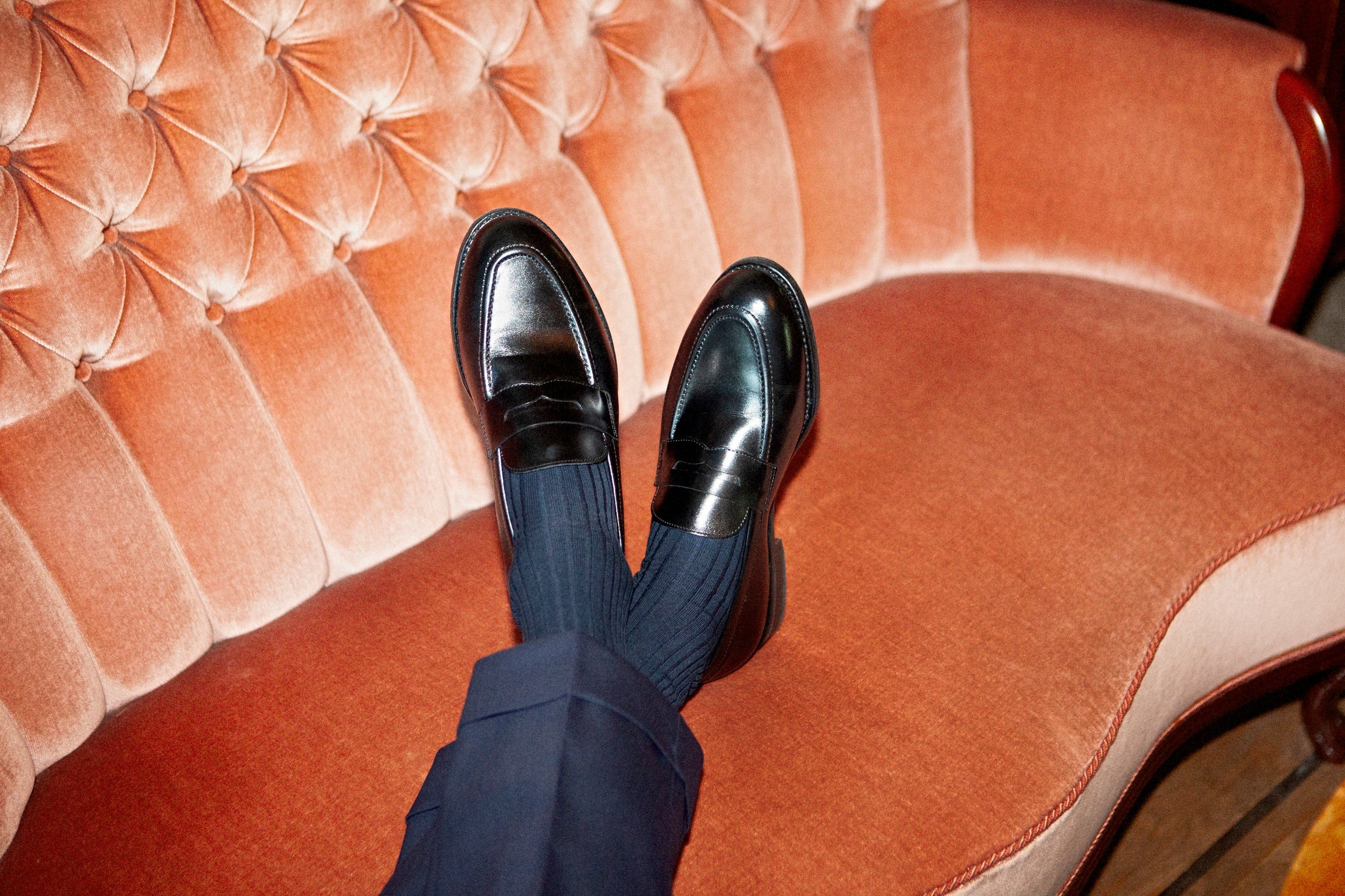 Black penny loafers worn with navy socks, feet crossed on a pink velvet sofa, showing the heel and collar of the shoe.