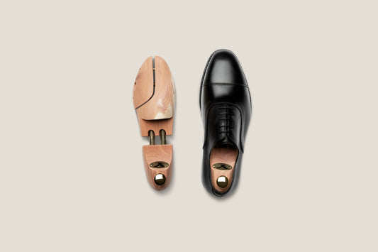 A single black oxford beside a wooden cedar shoe tree, viewed from above on a soft beige background.