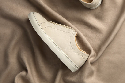 A pair of beige nubuck Alnö II sneakers arranged on draped tan satin fabric under warm side light.