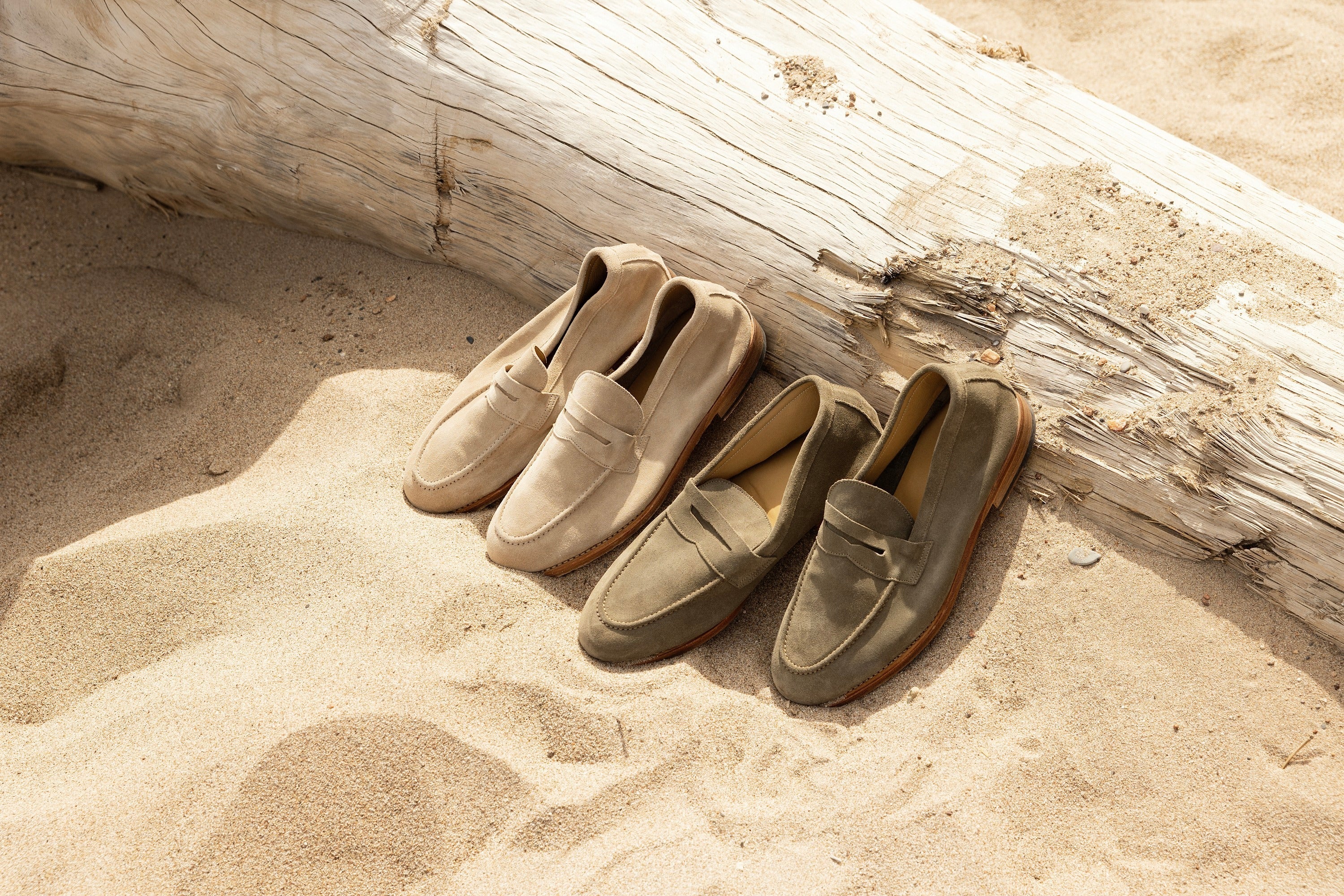 Two pairs of suede loafers, one sand and one olive, resting on sand beside a weathered driftwood log.