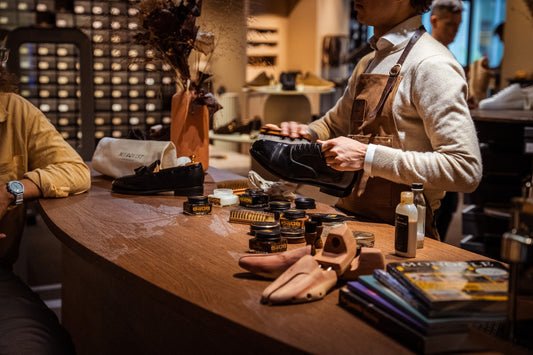 An aproned artisan applies polish to a black oxford at a wooden shine-bar counter, with open tins of wax and a horsehair brush nearby under warm light.