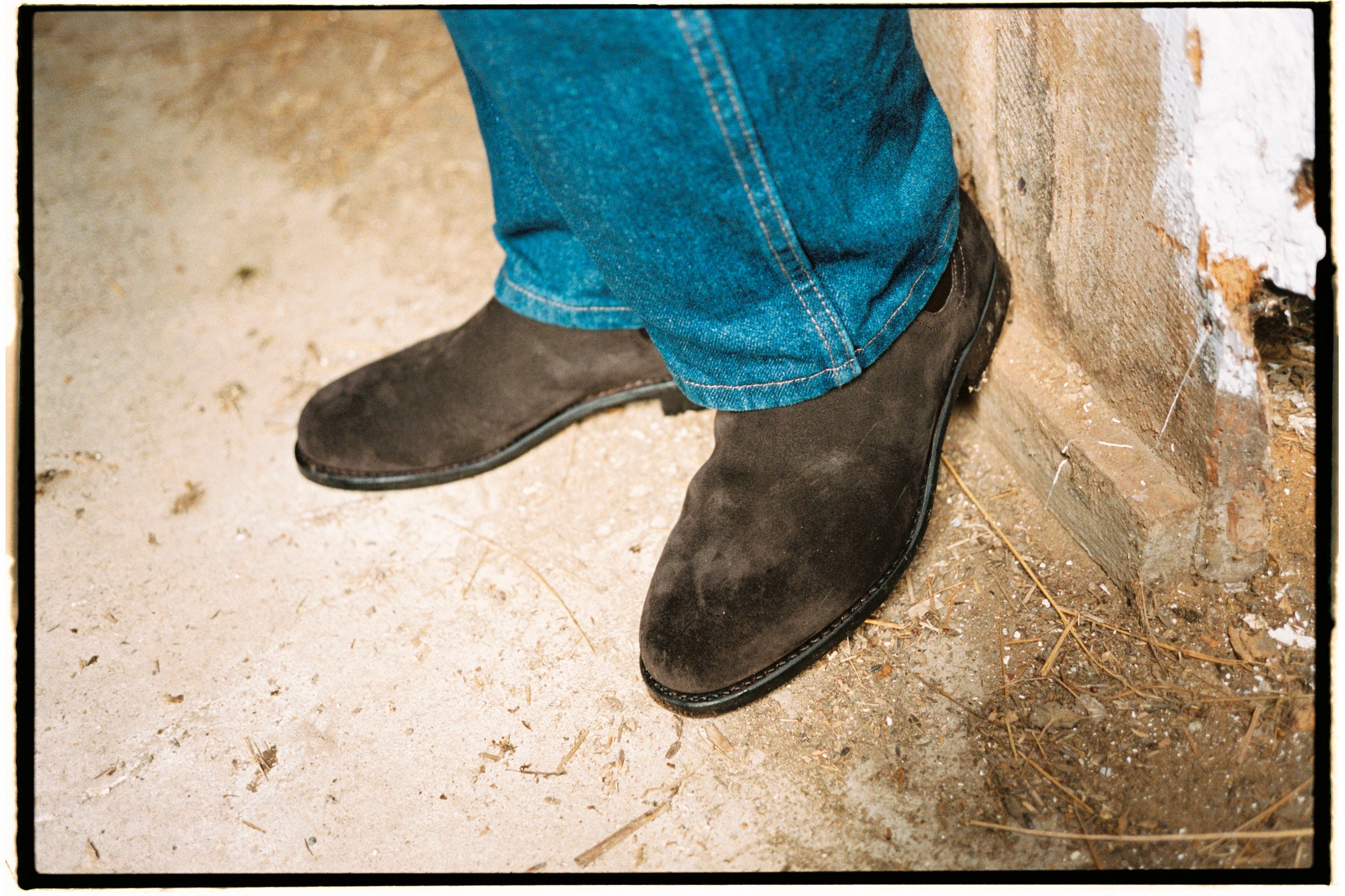 Dark brown suede Chelsea boots worn with blue jeans, standing on a concrete floor beside a whitewashed wooden wall in warm daylight.