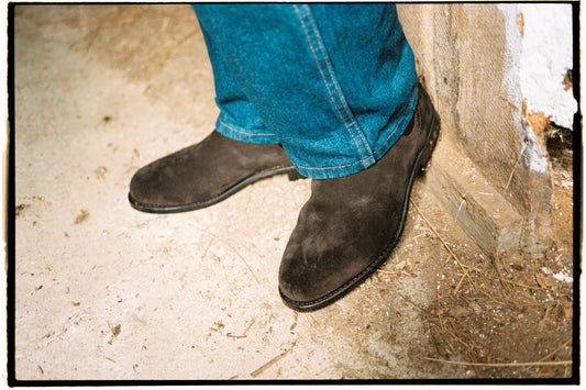 Dark brown suede Chelsea boots worn with blue jeans, standing on a concrete floor beside a whitewashed wooden wall in warm daylight.
