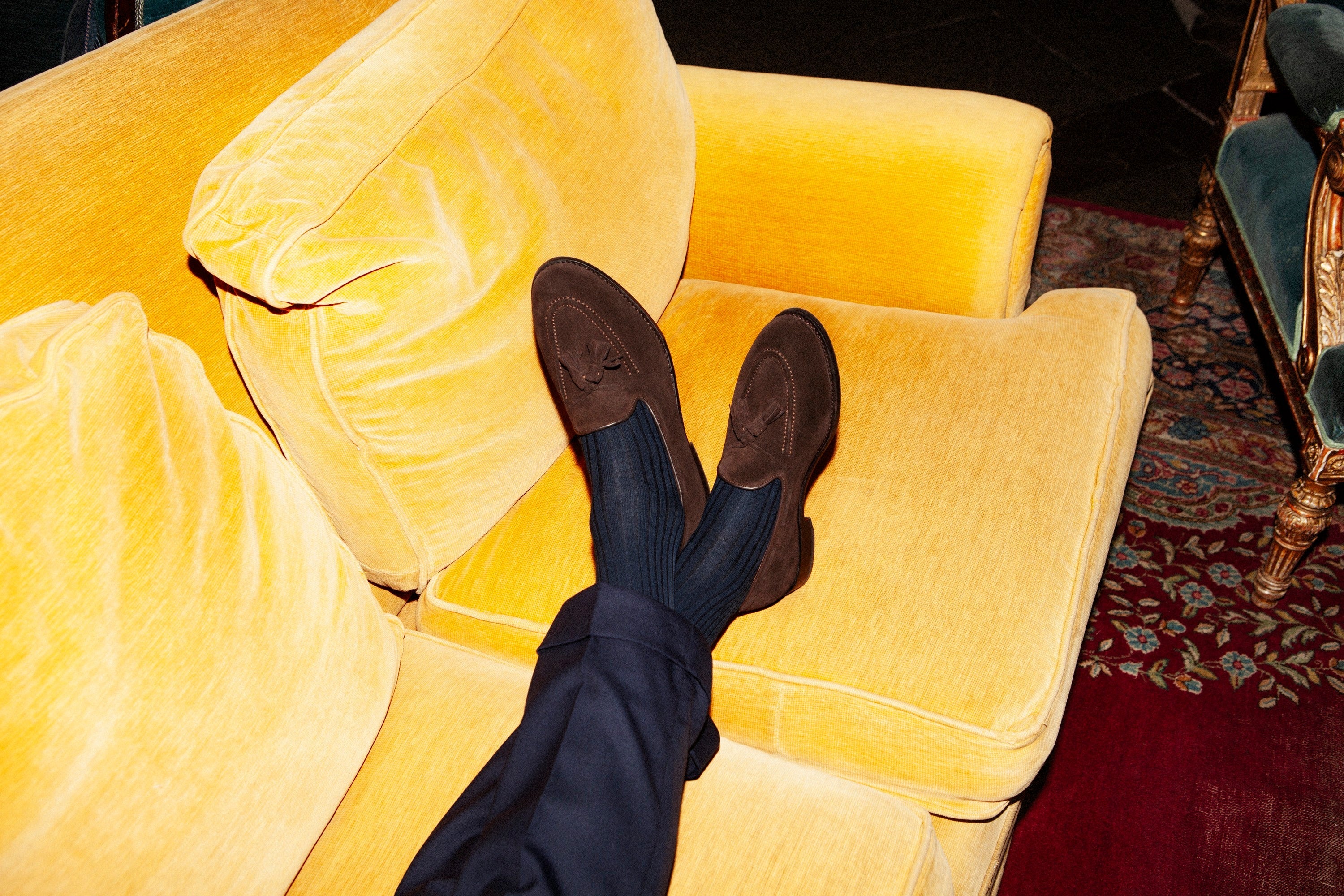Dark-brown suede horsebit loafers with navy trousers, feet crossed on a golden-yellow velvet sofa.