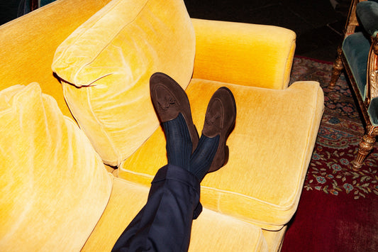 Dark-brown suede horsebit loafers with navy trousers, feet crossed on a golden-yellow velvet sofa.