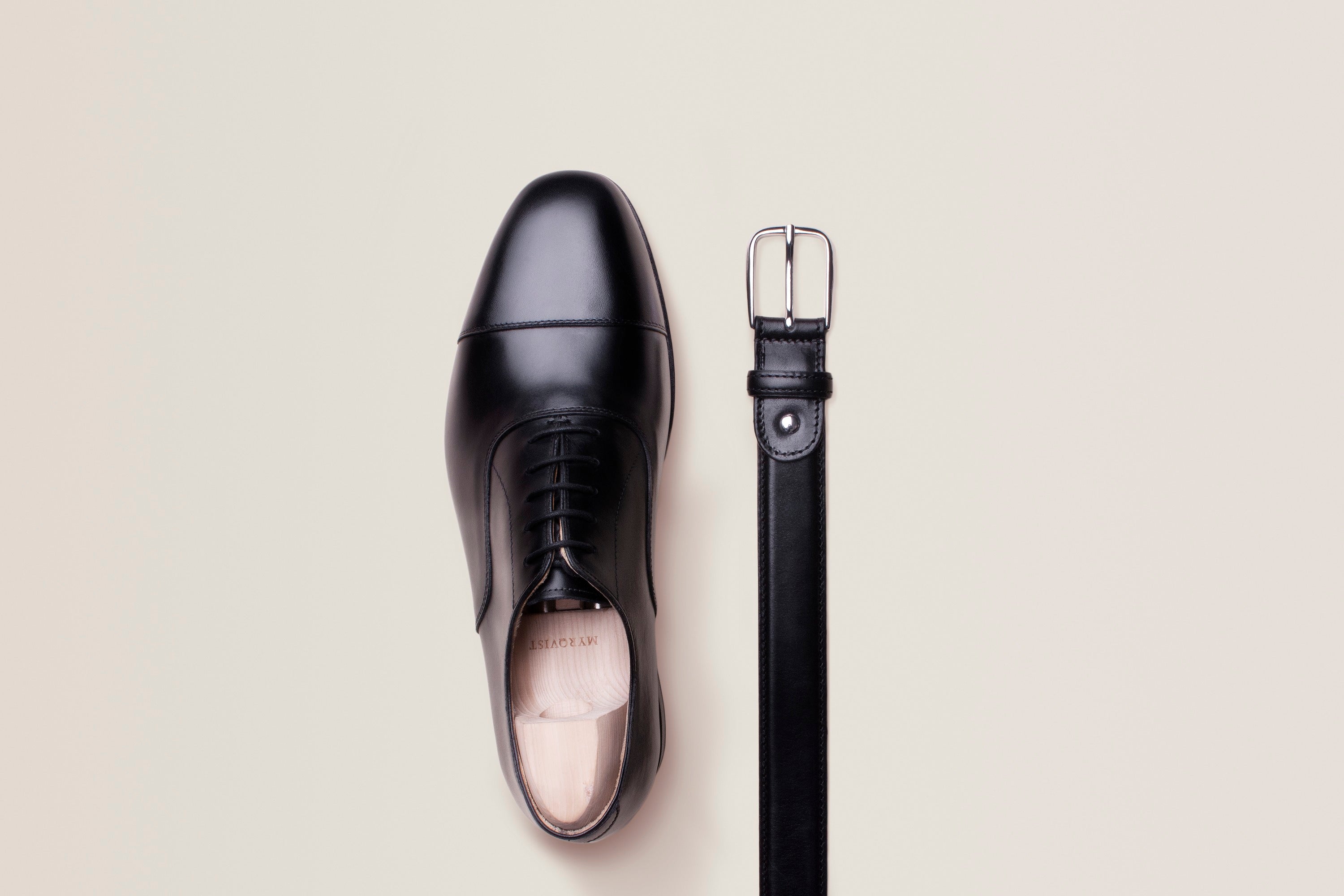 A black cap-toe leather oxford shoe lying beside a rolled black leather belt on a pale neutral background.