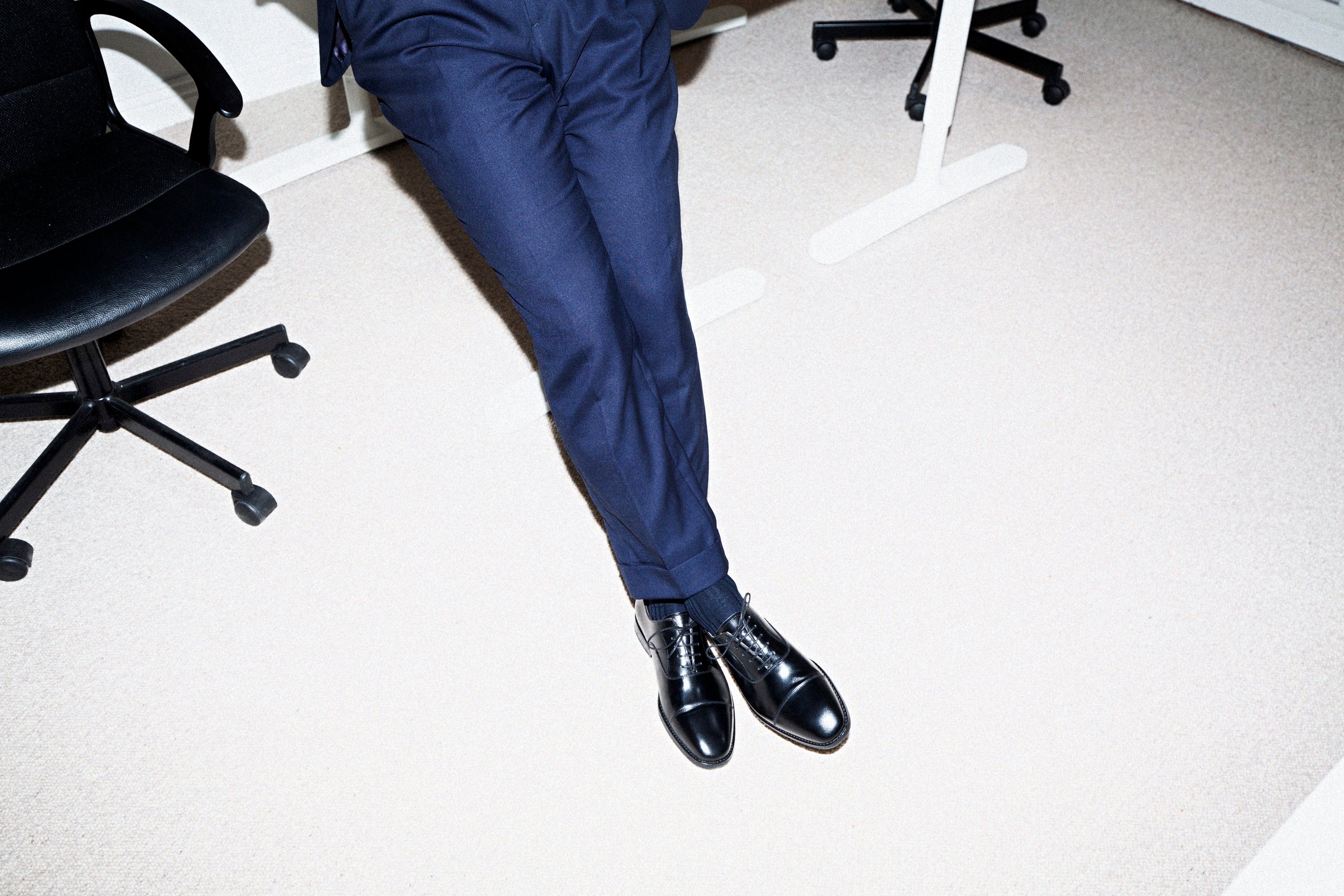 A man's black cap-toe oxford shoes with closed lacing, worn with navy suit trousers in an office.