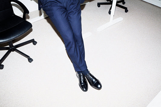 A man's black cap-toe oxford shoes with closed lacing, worn with navy suit trousers in an office.