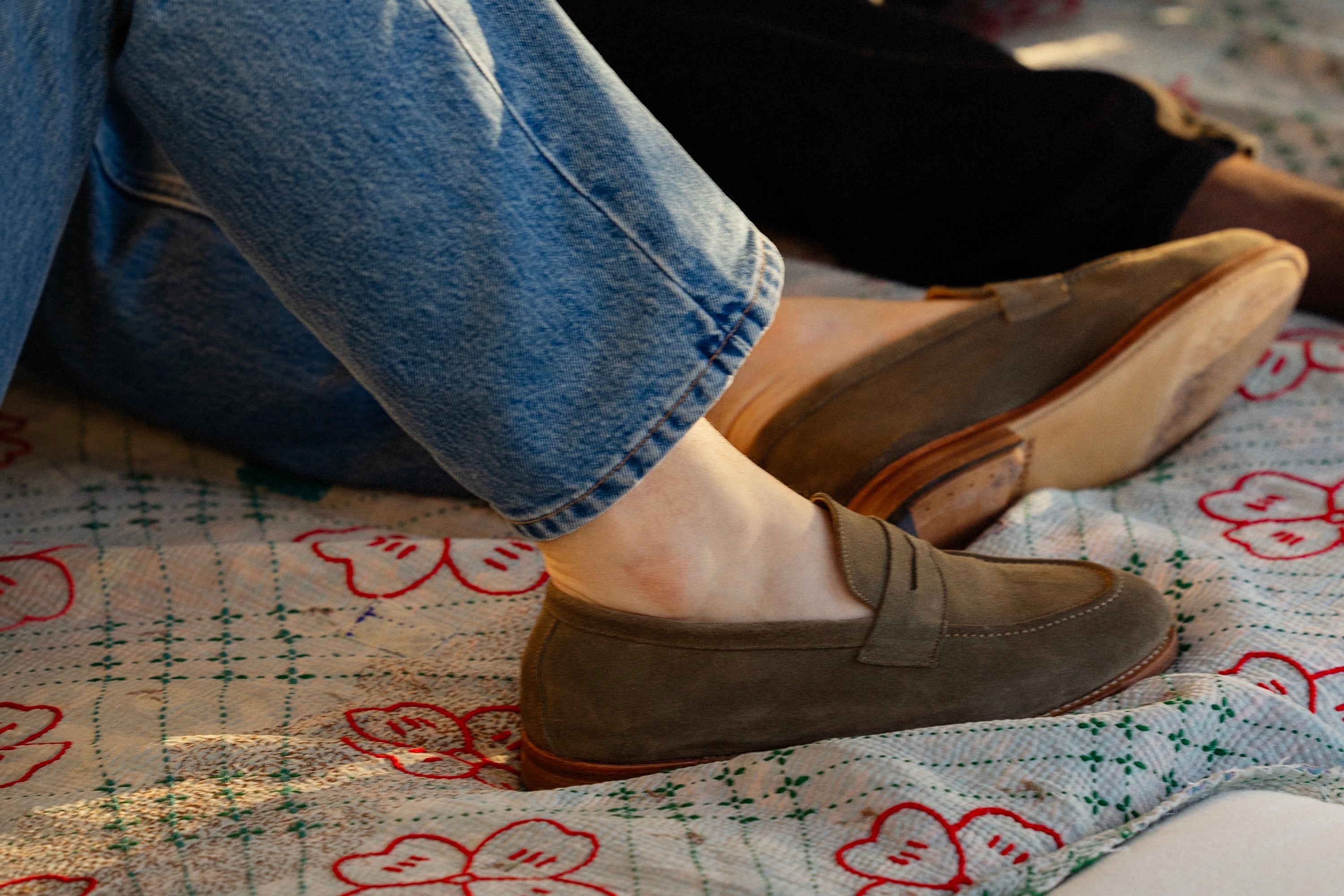 A brown suede penny loafer worn without socks, paired with rolled-up jeans.