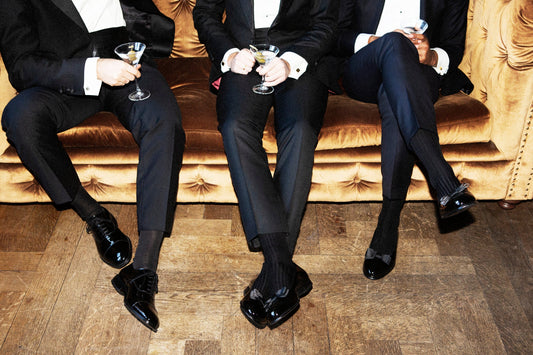 Three seated men in black tie holding cocktails, their patent oxfords and dark socks in the foreground on a wooden floor.