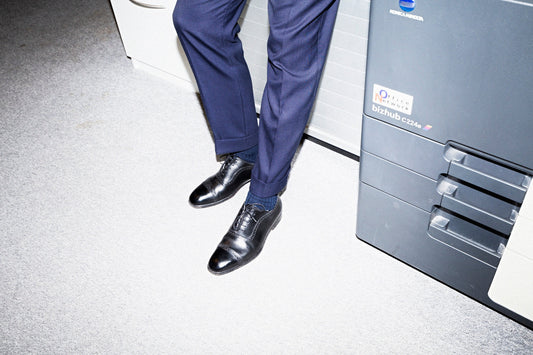 A man in navy suit trousers seated on the edge of an office copier, one black cap-toe oxford shoe pointed toward the camera.