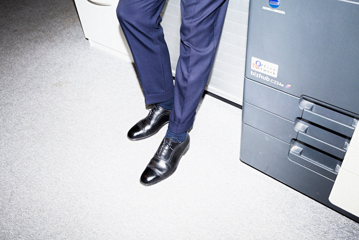 A man in navy suit trousers seated on the edge of an office copier, one black cap-toe oxford shoe pointed toward the camera.