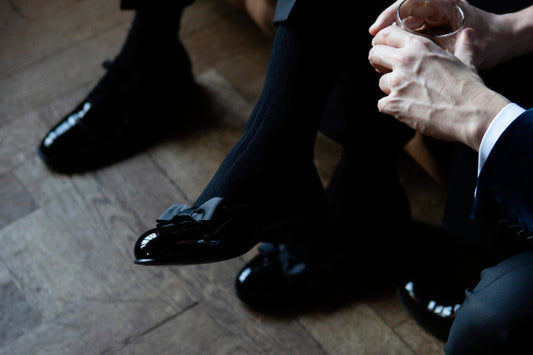 Crossed legs in a tuxedo with glossy black patent shoes on a parquet floor, a hand holding a drink to one side, in soft window light.