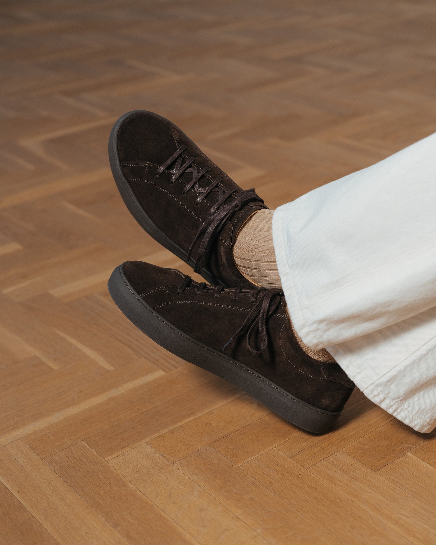 Oaxen Monochrome Wildledersneaker in Dunkelbraun, getragen mit heller Stoffhose und beigen Socken auf Holzboden, Ansicht von oben zeigt die monochrome Low-Top-Silhouette im Alltag.