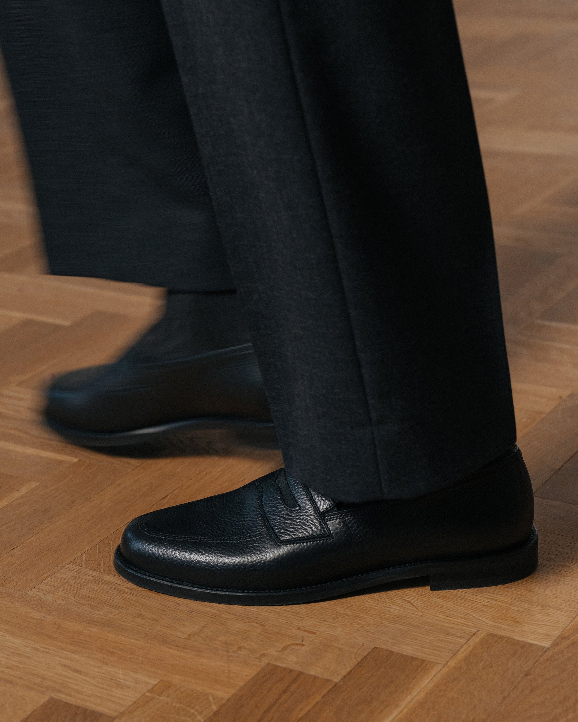 Sigtuna – Black Natural Grain loafers worn with dark grey trousers on a wooden parquet floor.