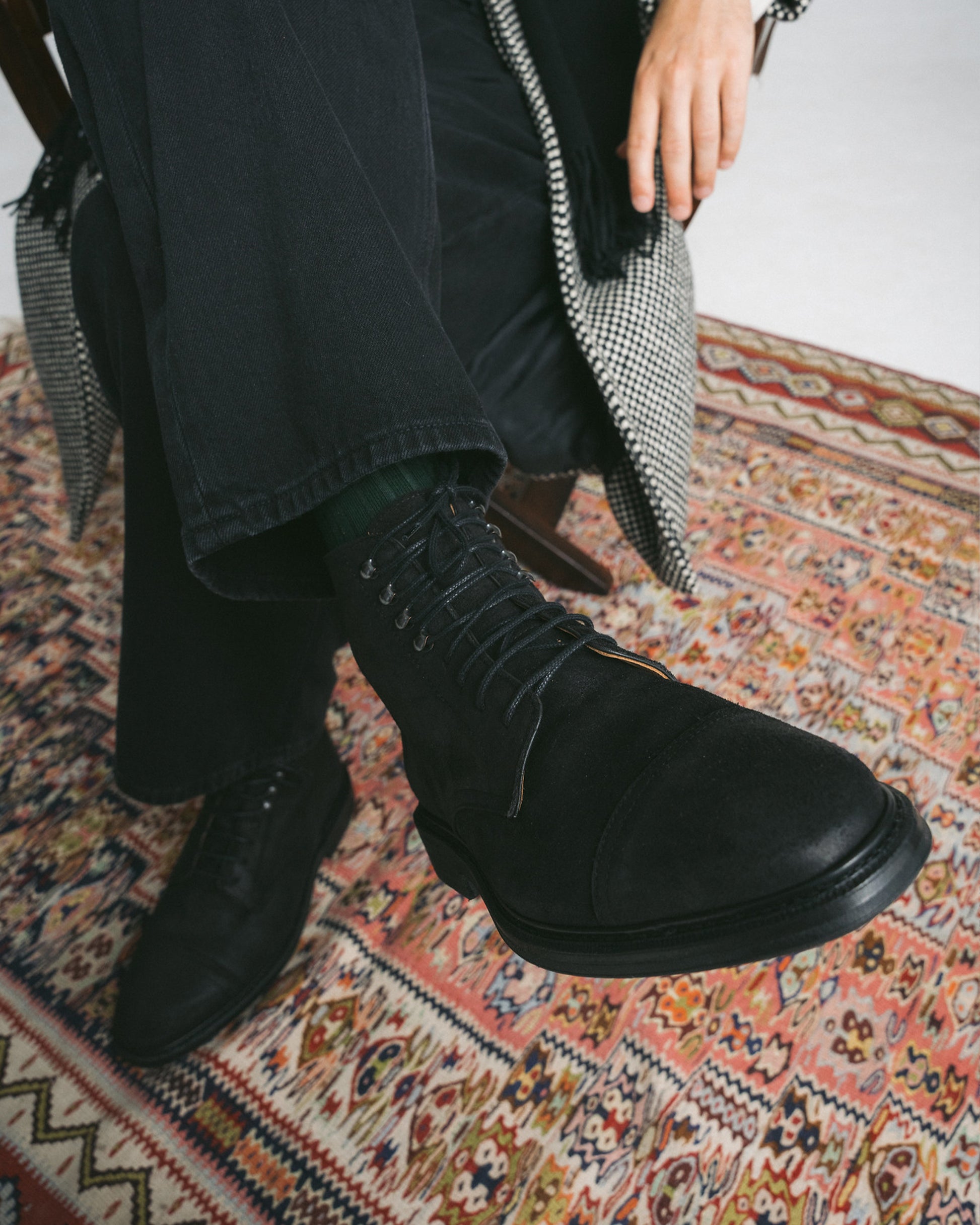 A person sitting on a chair wearing Abisko – Black Waxed Suede boots with green socks and black trousers.