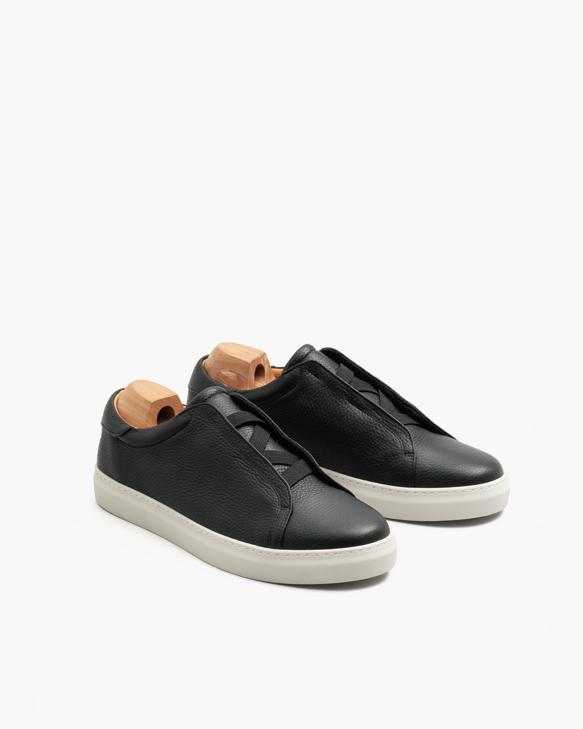 Pair of Alnö II slip-on sneakers in black natural grain leather with a minimalist design, subtle stitching, and white rubber soles, shown with wooden shoe trees inserted.