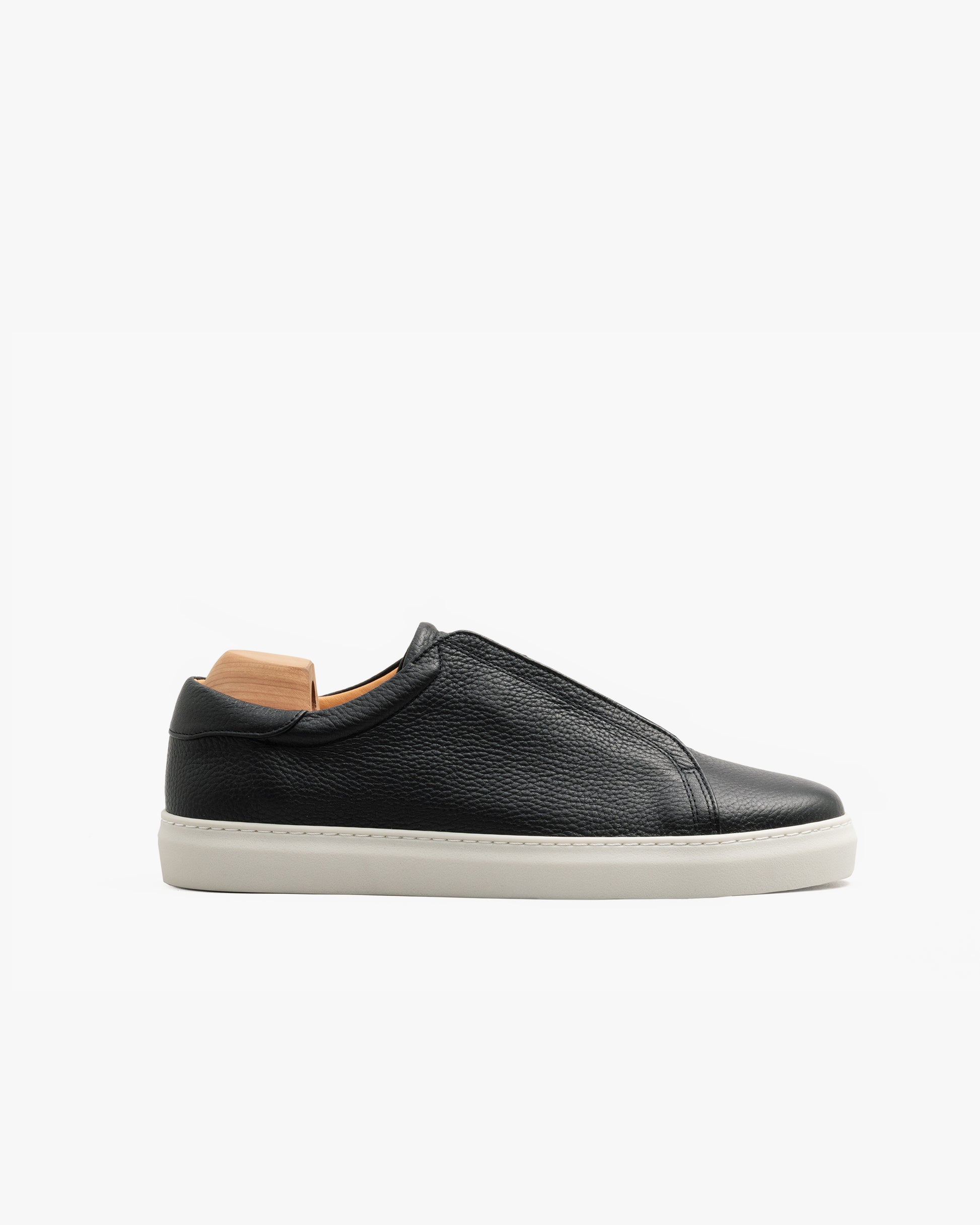 Low-top slip-on sneaker in black natural grain leather with clean lines, subtle stitching, and a contrasting white rubber sole, shown in profile with a wooden shoe tree inside.