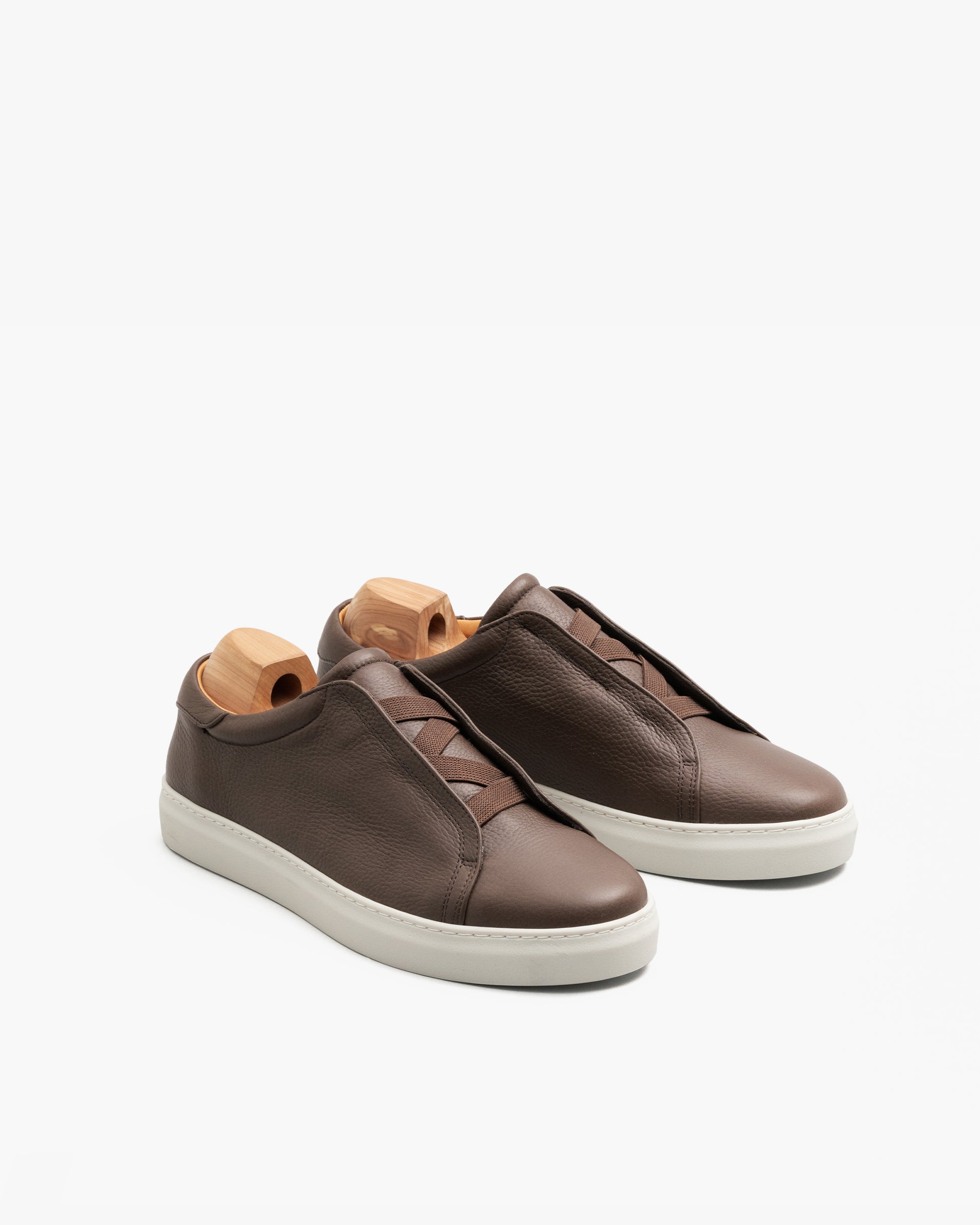 Pair of Alnö II sneakers in brown natural grain leather with a minimalist low-top design, partially concealed laces, and contrasting white cup soles, displayed with wooden shoe trees on a clean white background.