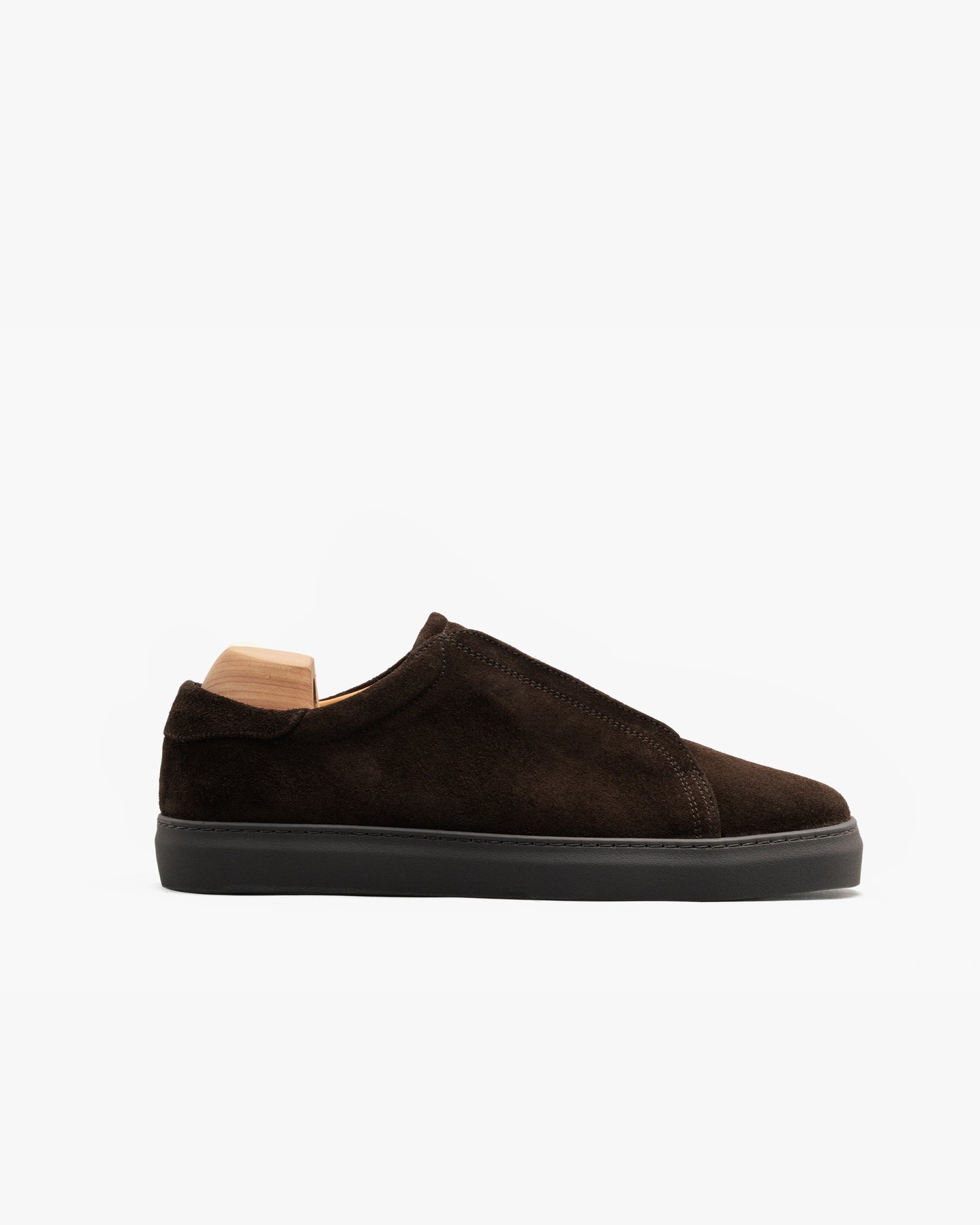 Dark brown suede slip-on sneaker with a monochrome design, featuring clean uninterrupted lines, tonal stitching, and a color-matched rubber sole, shown in profile with a wooden shoe tree inside.